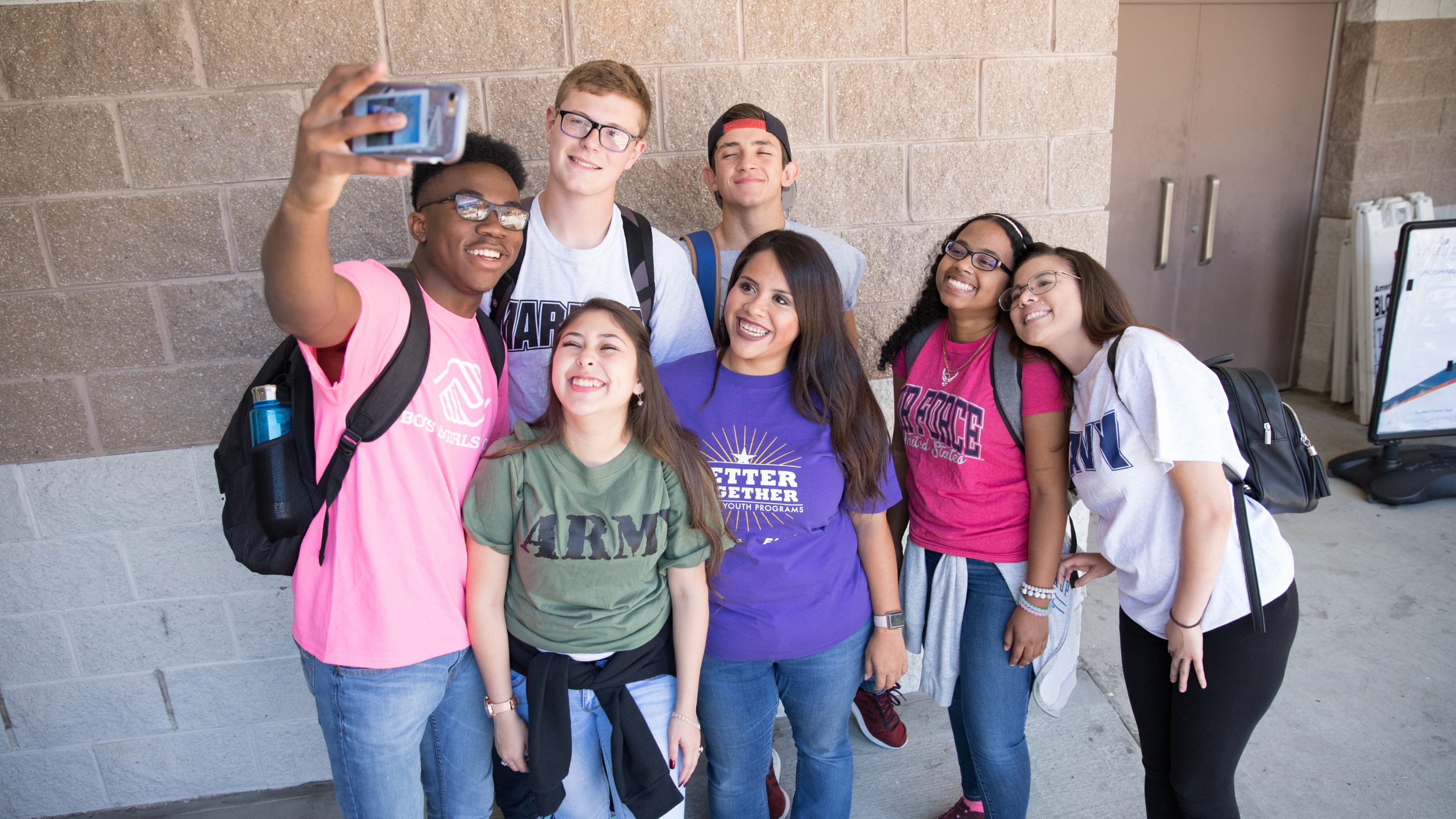Group of seven diverse teenagers taking a selfie together outdoors by a brick wall, smiling and dressed casually with backpacks.