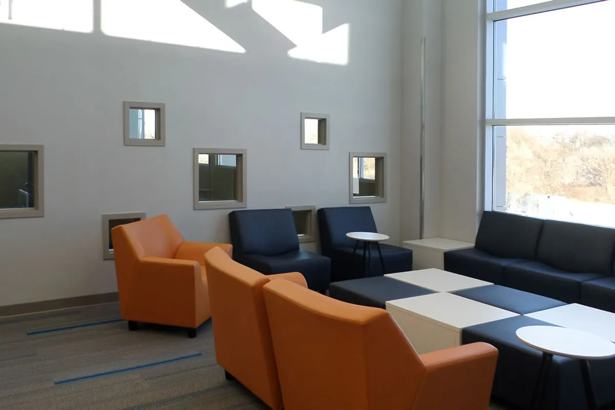 Modern lounge area with orange and black armchairs, black sofa, small white tables, large window, and decorative square windows on the wall.