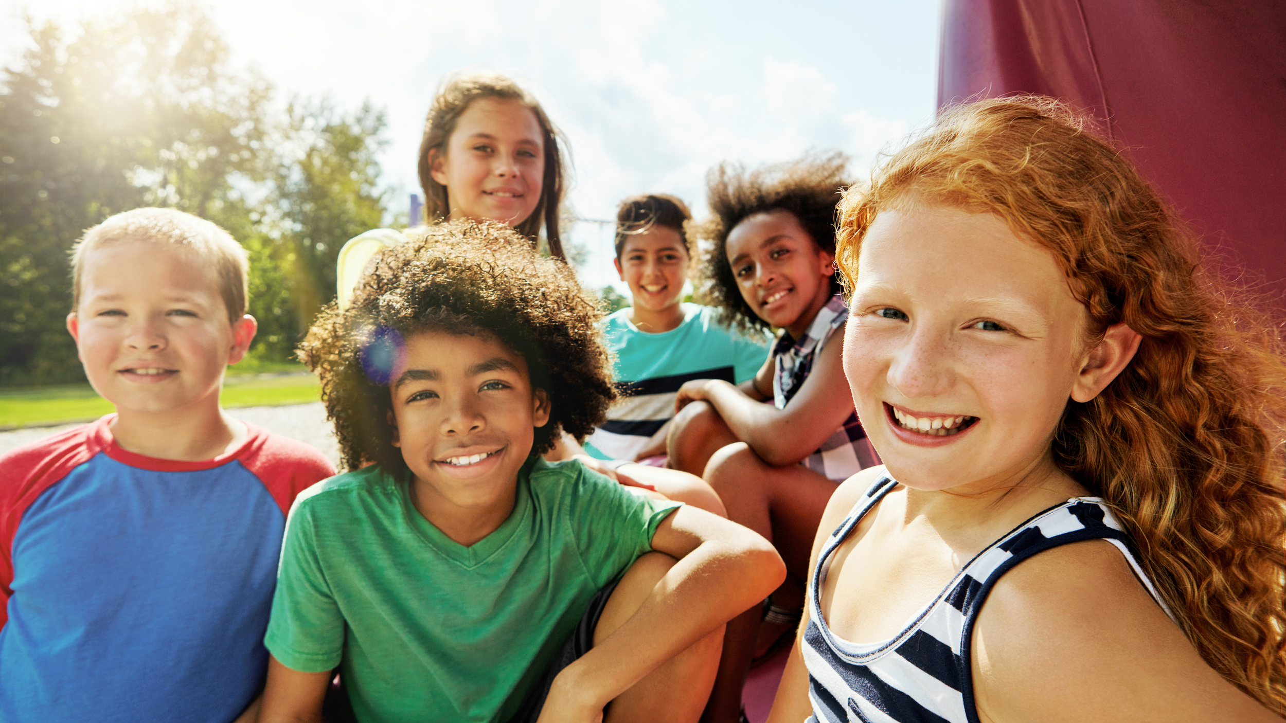 Six children smiling and sitting outdoors on a sunny day.