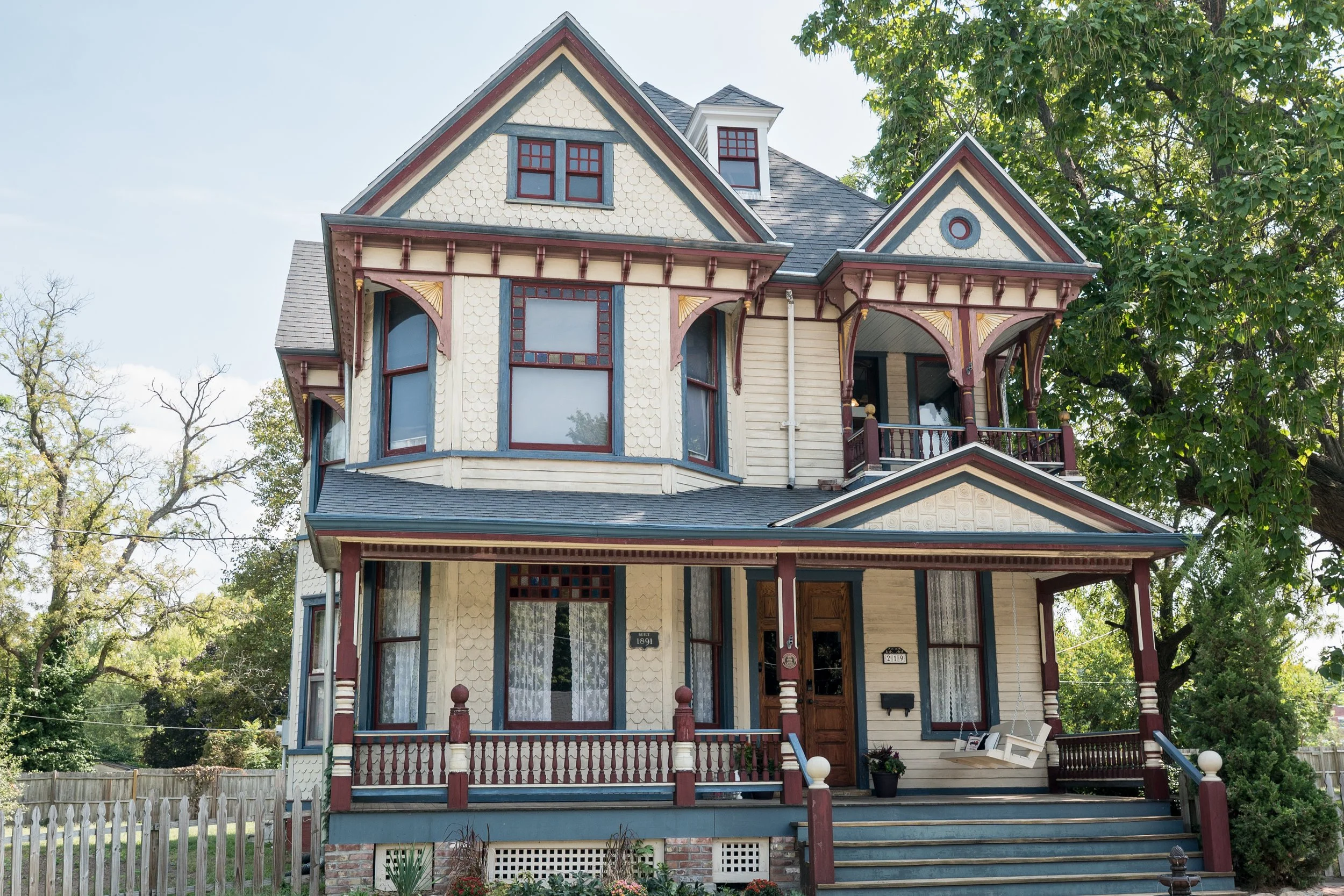 A Victorian style house with ornate detailing, decorative trim, a front porch, large bay window, and a gabled roof surrounded by trees.