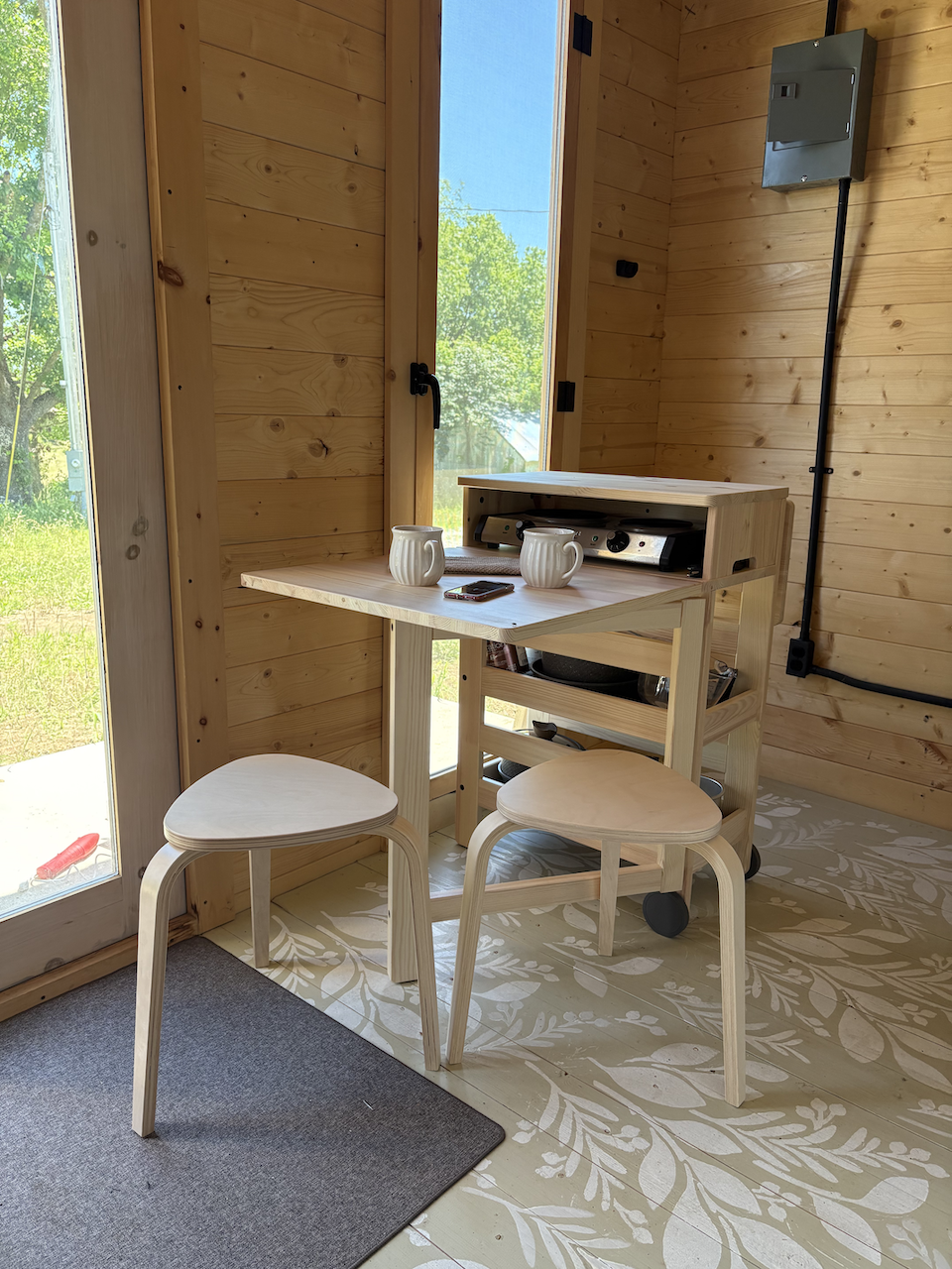Interior of a wooden room with a small wooden kitchen cart, two beige wooden stools, two white mugs, and a smartphone on the cart. A sliding glass door with a view of green trees outside is visible. The room has wood paneling on the walls and a patterned floor.