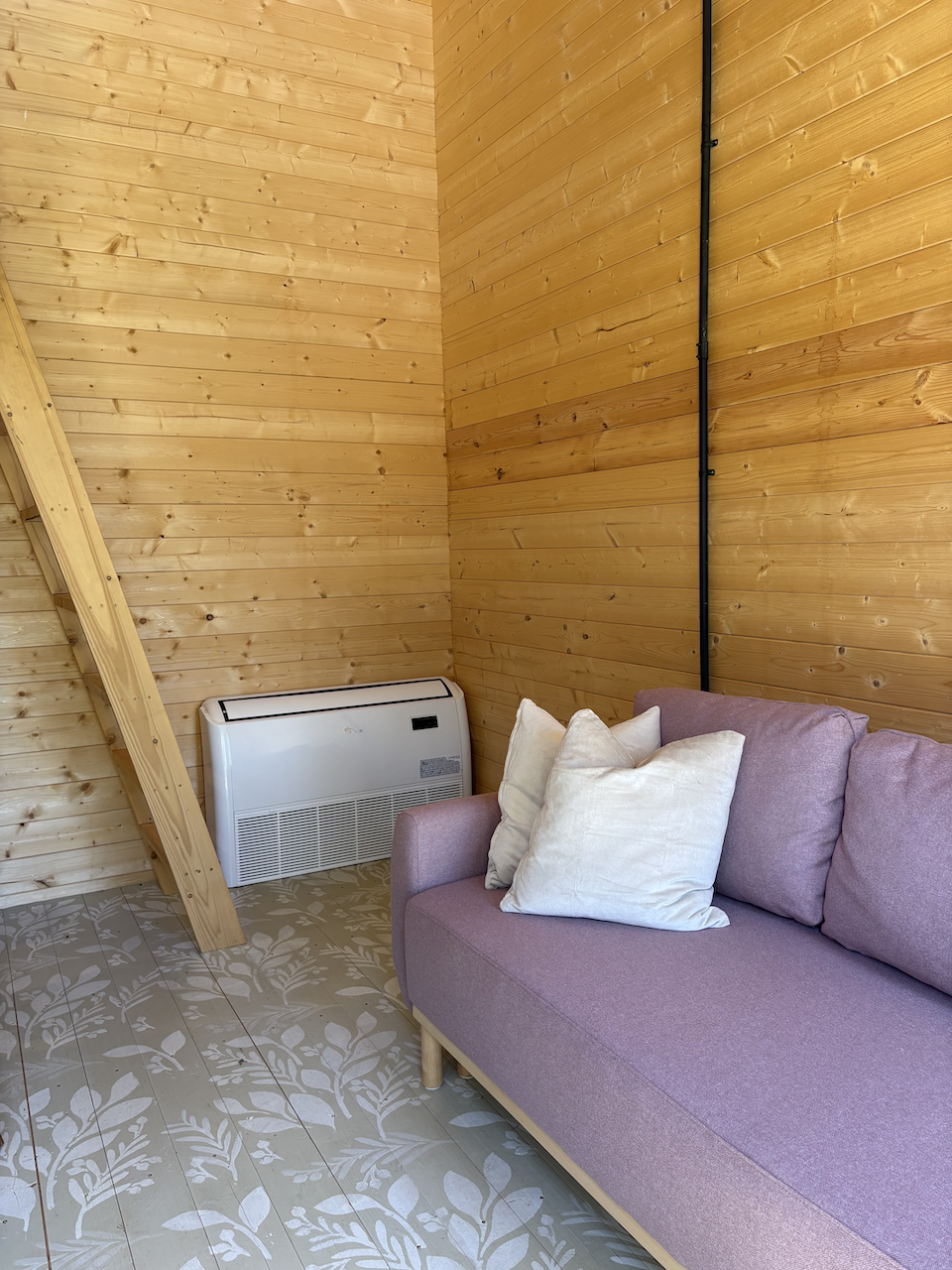 A cozy interior corner with light purple sofa with white throw pillows, wooden wall paneling, patterned floor tiles, a heater, and a wooden staircase.