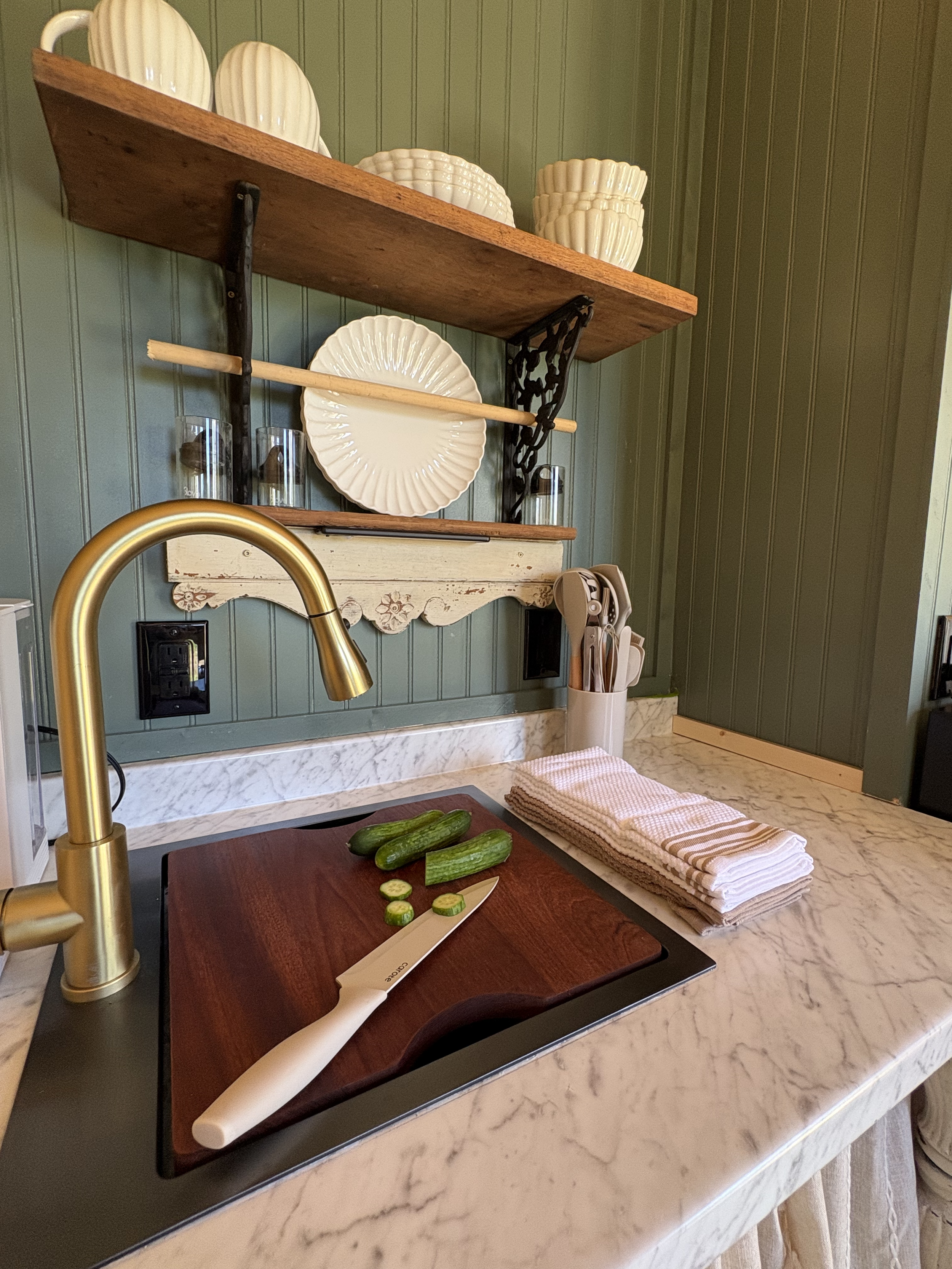 A kitchen with a marble countertop, a gold faucet, and a dark wooden cutting board with sliced cucumbers. A knife rests on the cutting board. In the background, there's a green paneled wall with open wooden and decorative ceramic shelves holding various dishes, bowls, and glasses. There is a stack of folded dish towels on the counter.