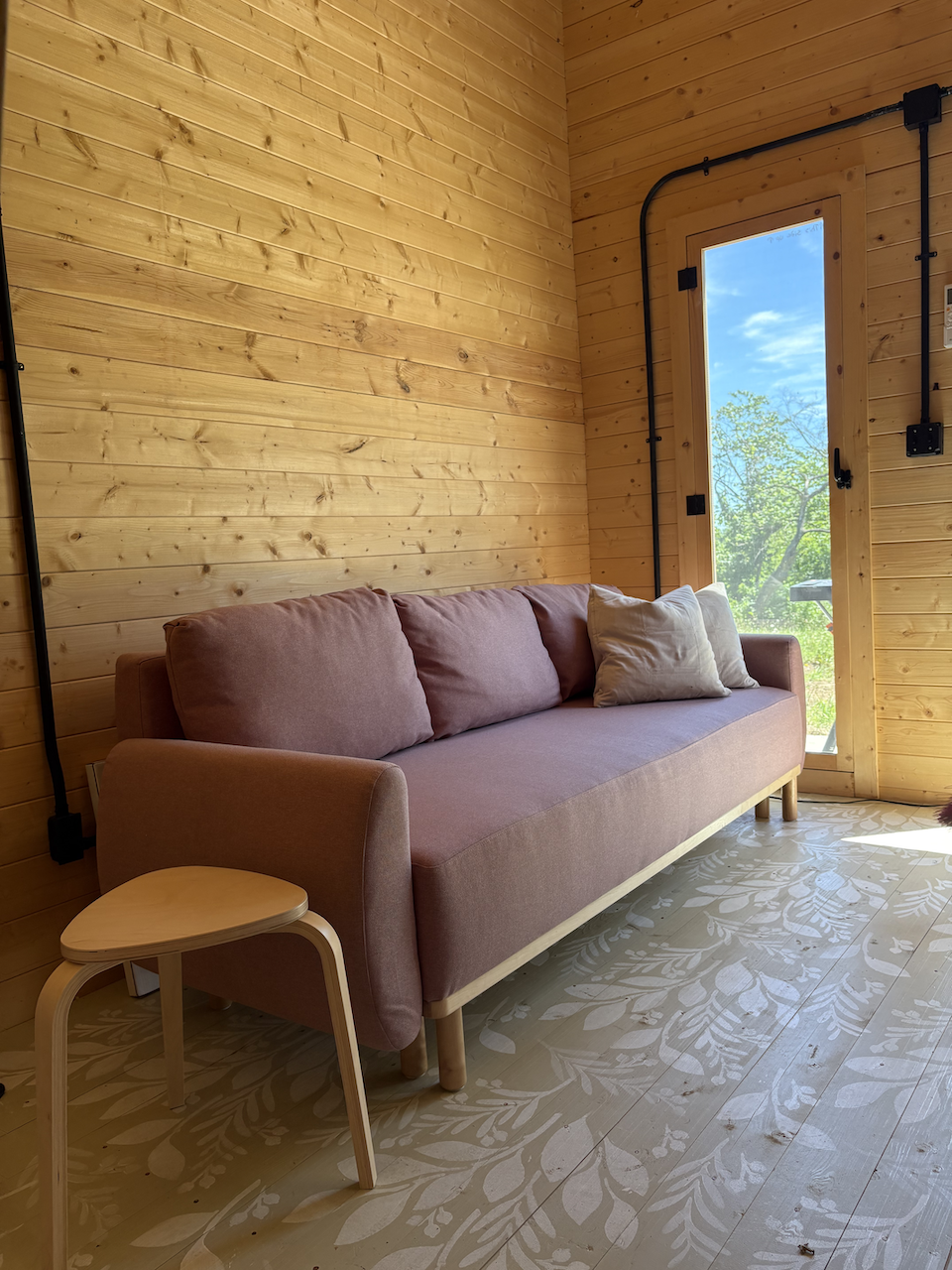 A cozy living room corner with a pink sofa, a small wooden side table, a light-colored pillow, and a large window showing a view of trees and blue sky, all set against wooden paneled walls and floor with a leaf pattern.
