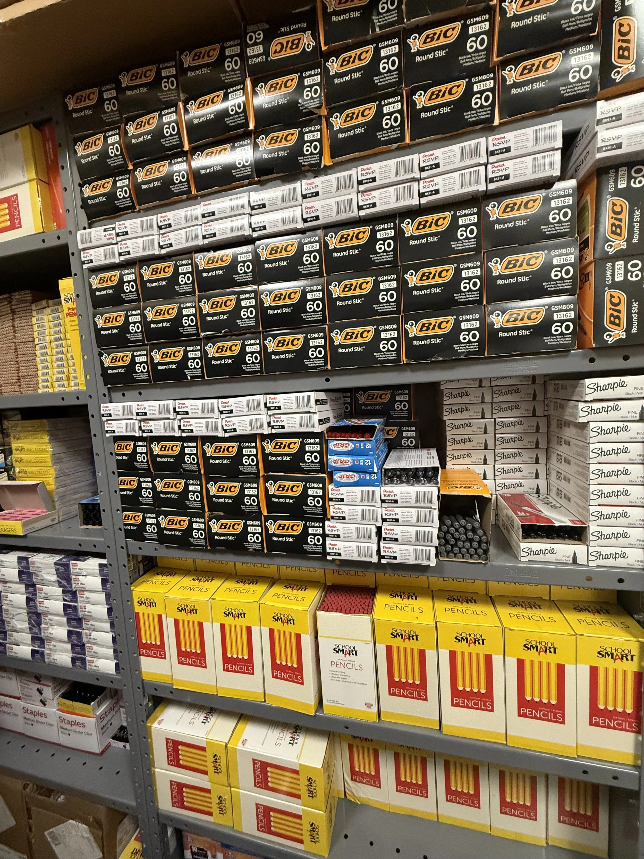 Shelf stocked with Bic round stick pens in black boxes, and larger boxes of school pencils in yellow and red packaging.
