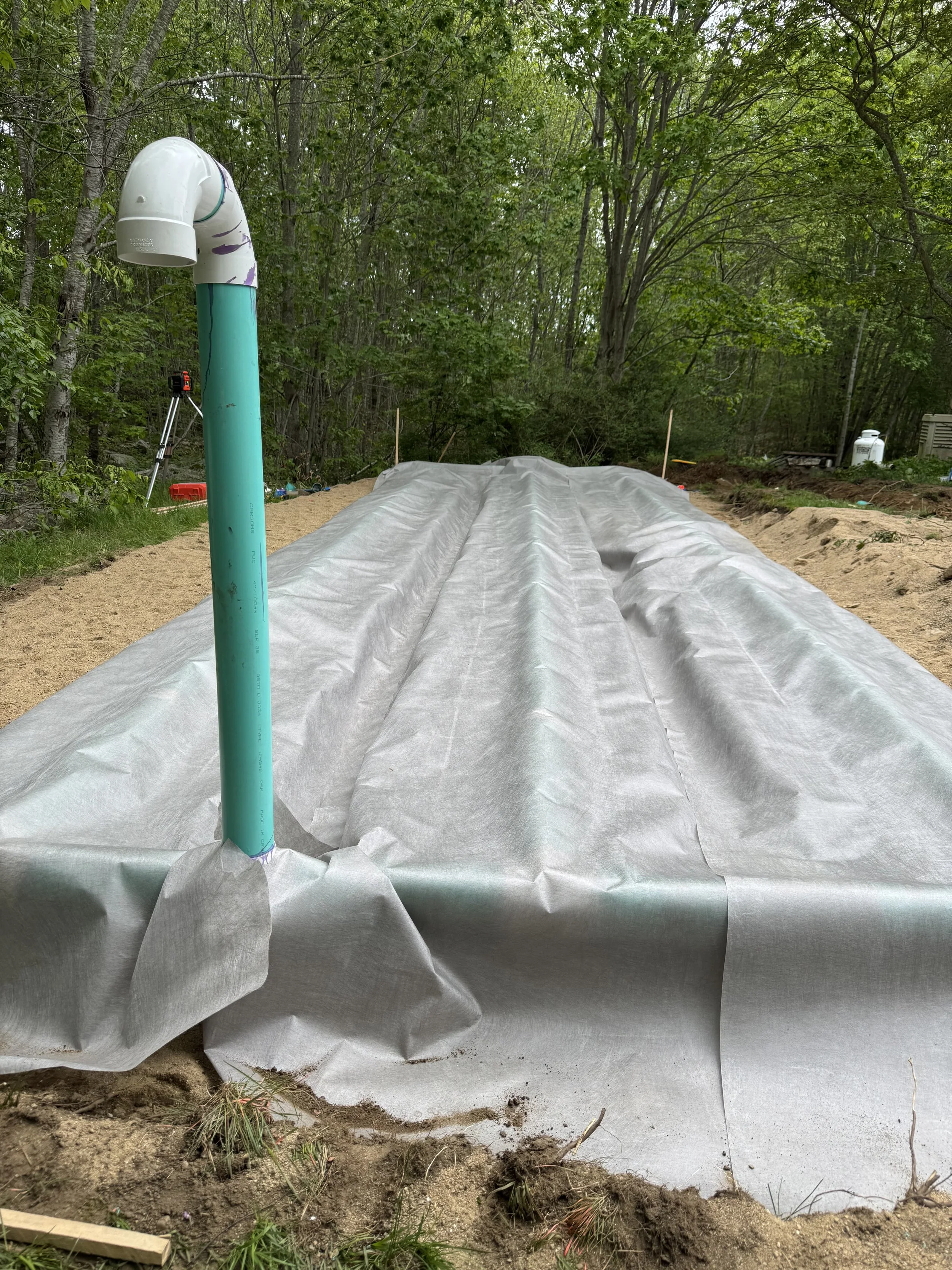 Green plastic piping protruding through a white fabric-covered trench at a construction or landscaping site, surrounded by trees and soil.