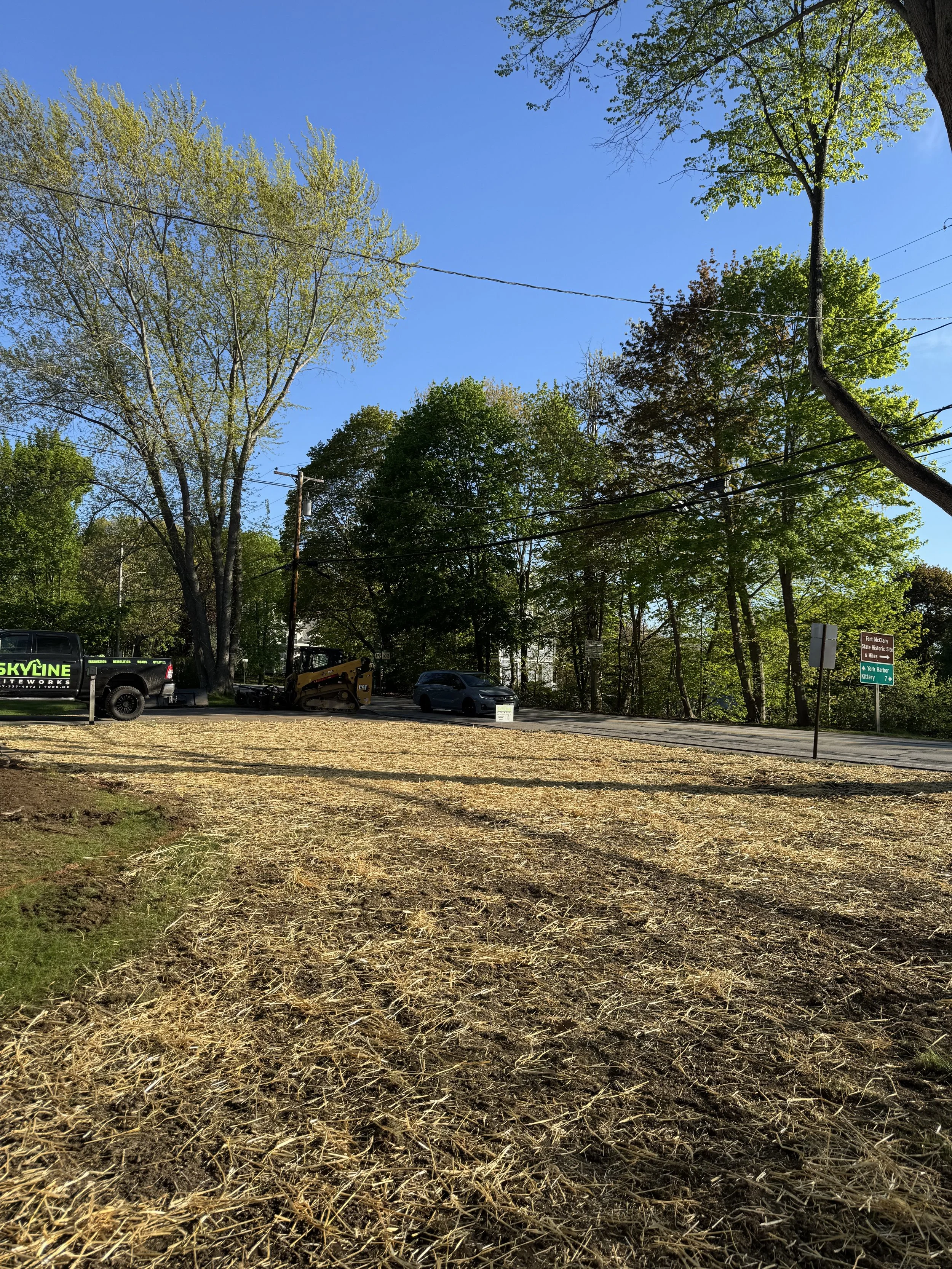 A street with trees on both sides under a clear blue sky. Construction vehicles and parked cars are visible along the road, and the ground in the foreground is covered with straw or dry grass.