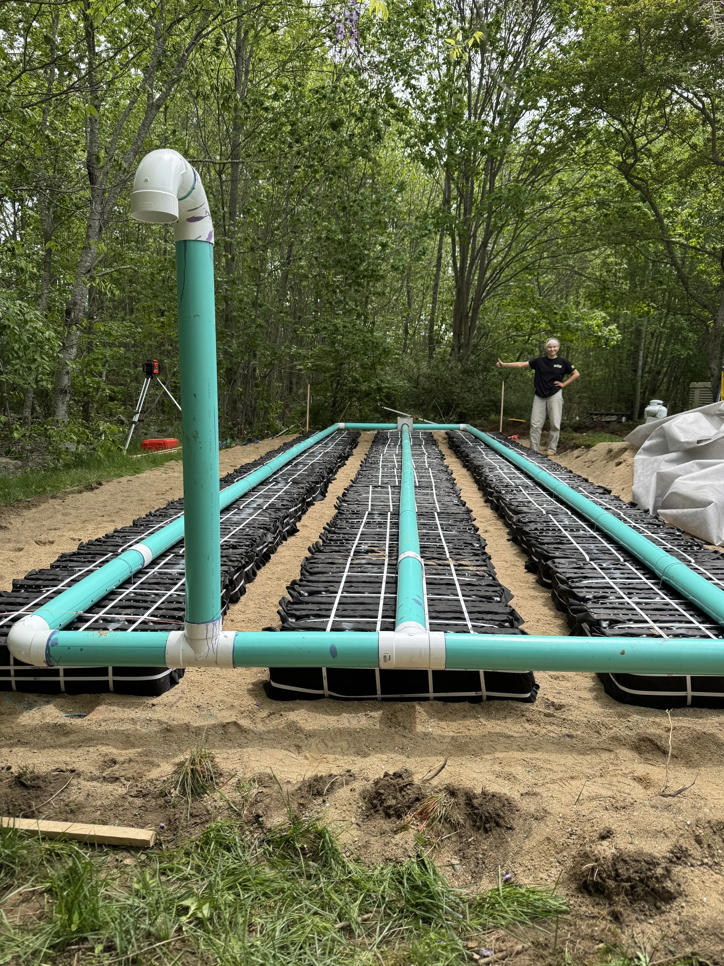 A person standing in a wooded area next to an underground irrigation system with exposed green pipes and black sub-surface drip emitters.