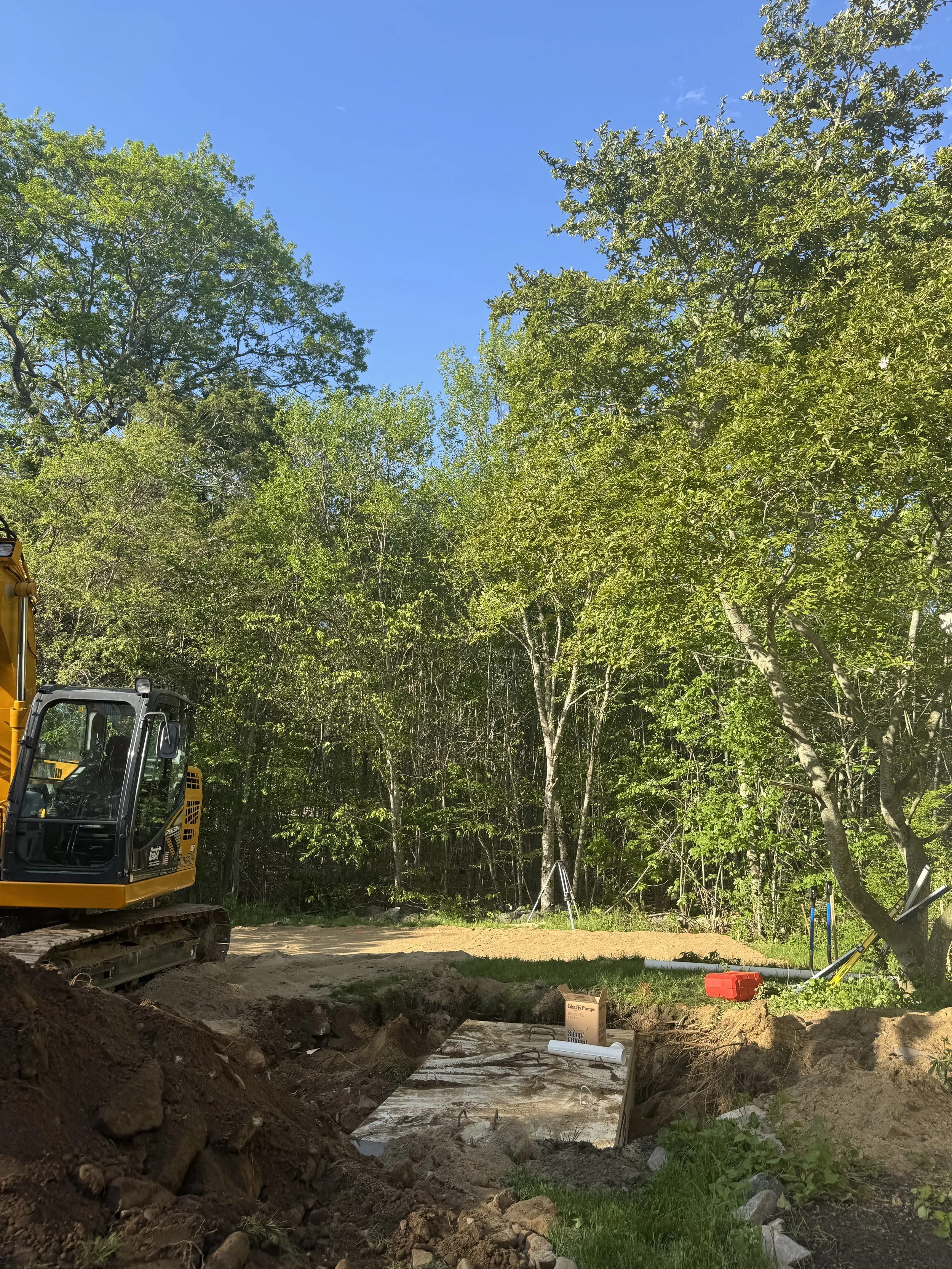 Construction site with a yellow excavator on the left, trees with green leaves on the right, and a clear blue sky in the background. There is a trench with concrete on the ground and construction tools near the trees.