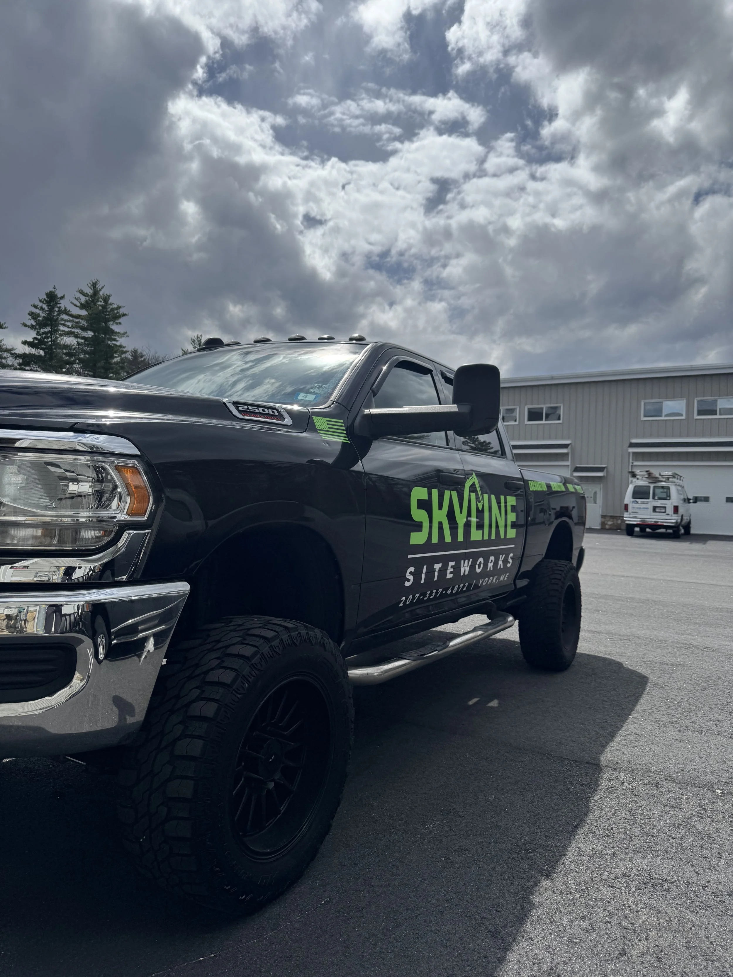 A black pickup truck with green and white company branding parked outside a large industrial building with white siding. The sky is partly cloudy.