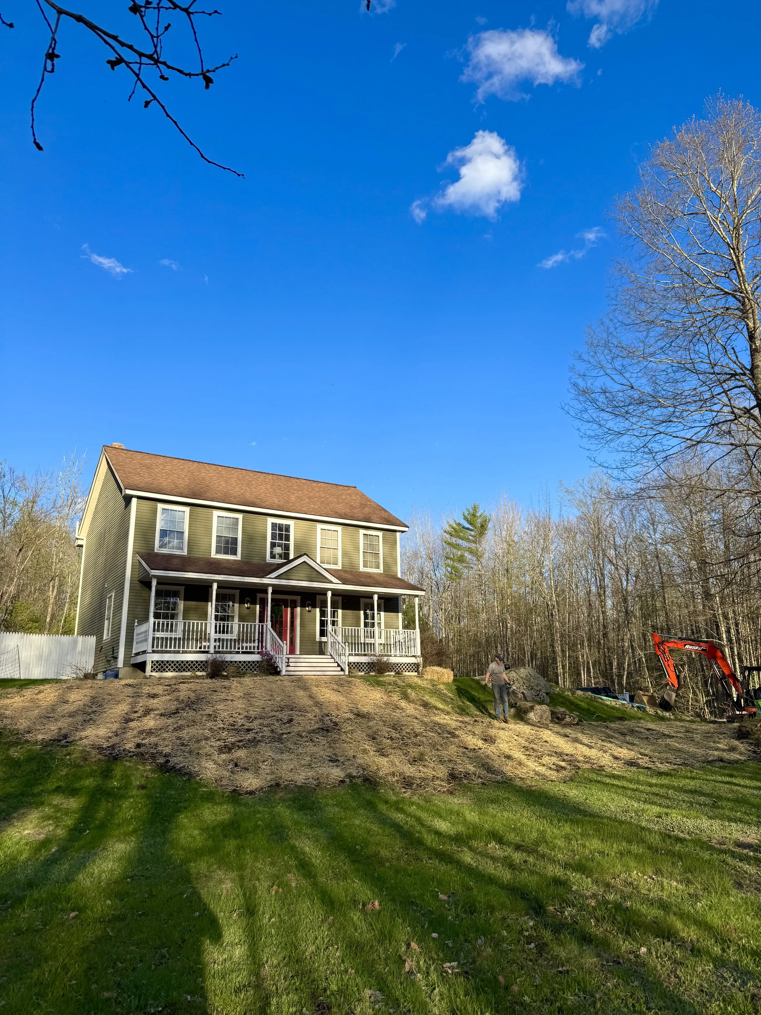 A two-story house with a porch and stairs, surrounded by a green lawn and trees, with a blue sky and clouds overhead. There is construction activity on the hillside to the right, with a person and an orange excavator at work.