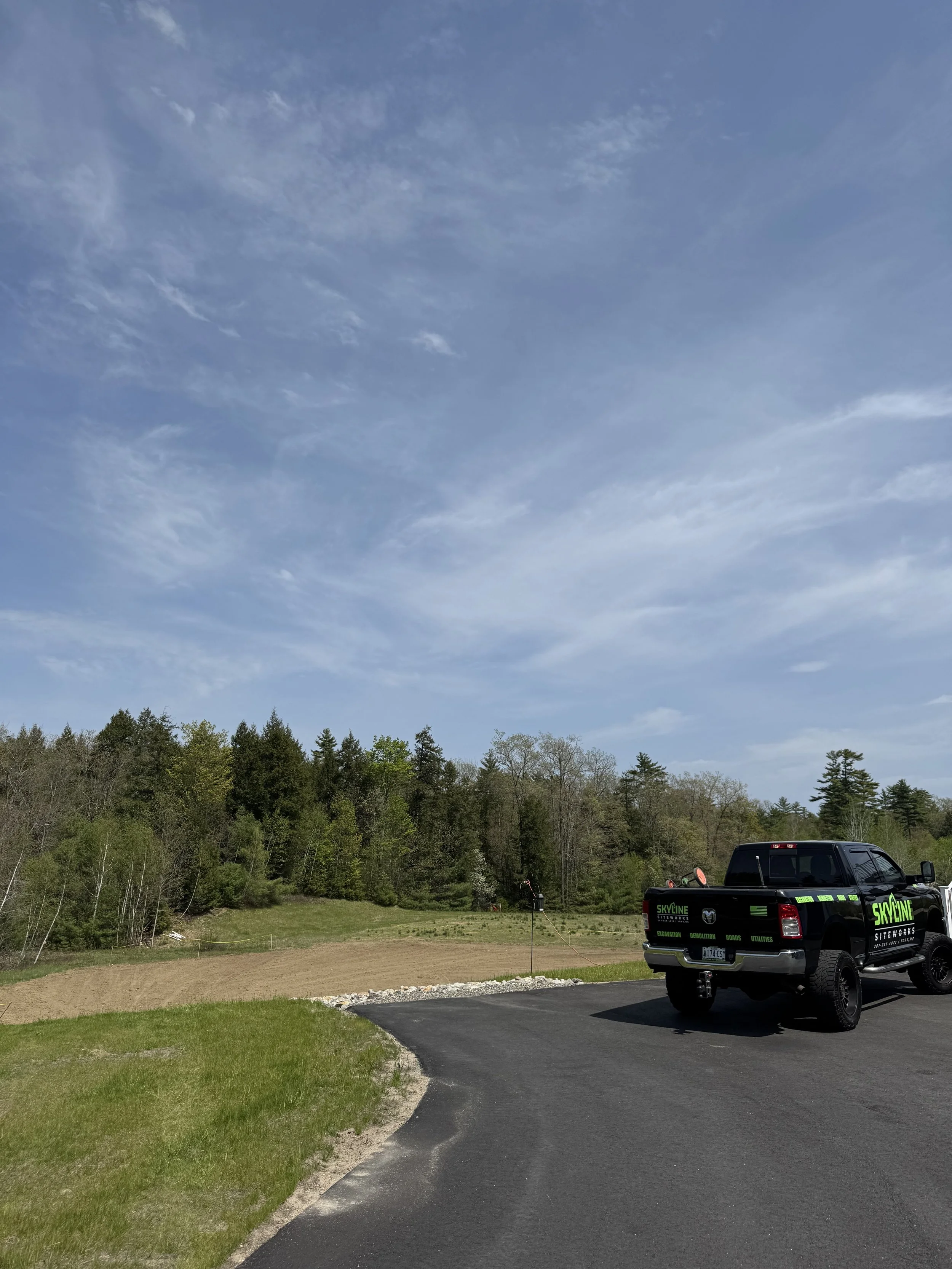 A black utility truck parked on a paved driveway next to a grassy area with trees in the background. The sky is partly cloudy with some blue visible.