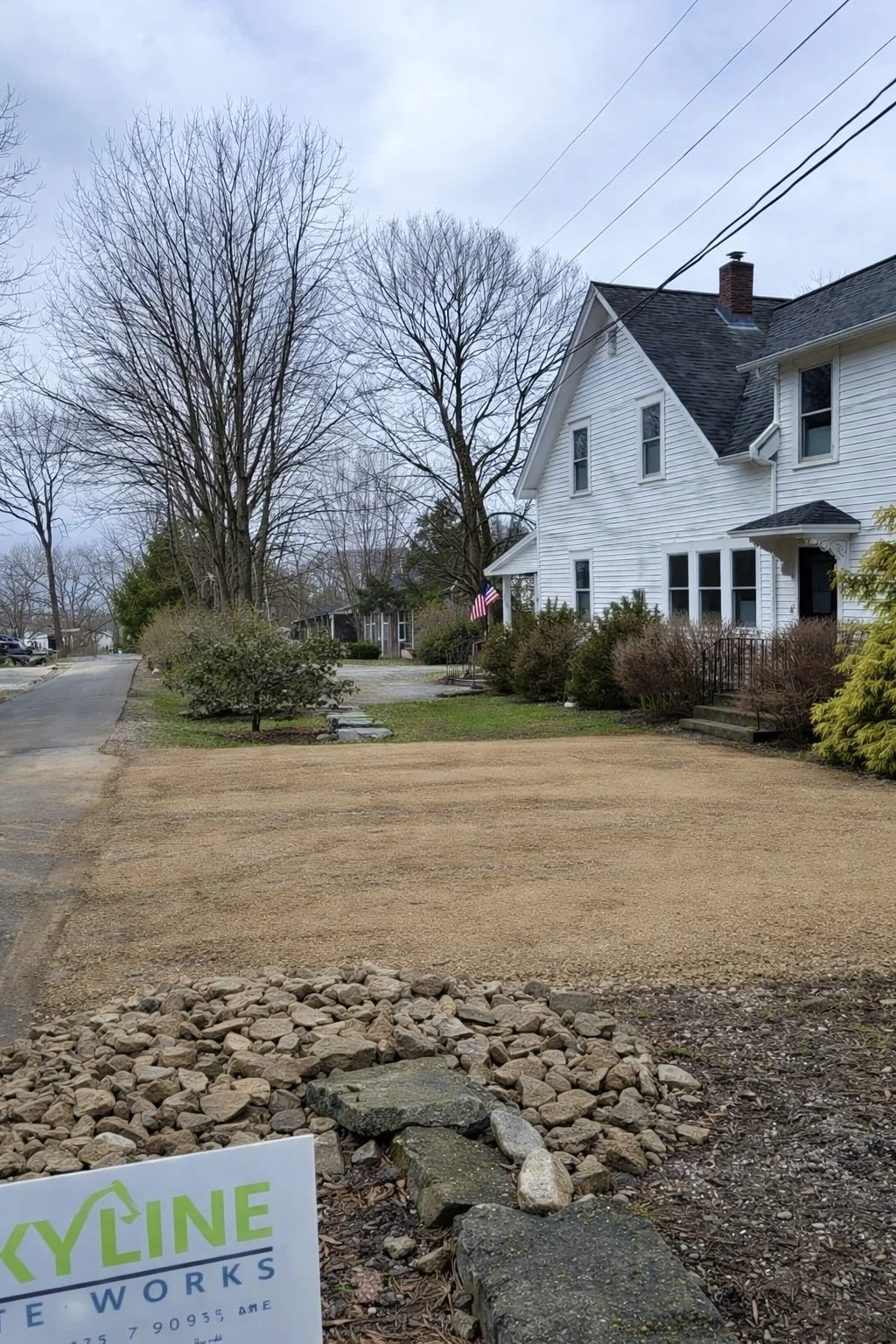 Residential street with a large white house, leafless trees, and a yard under construction with gravel, rocks, and a sign that reads 'Skyline'.