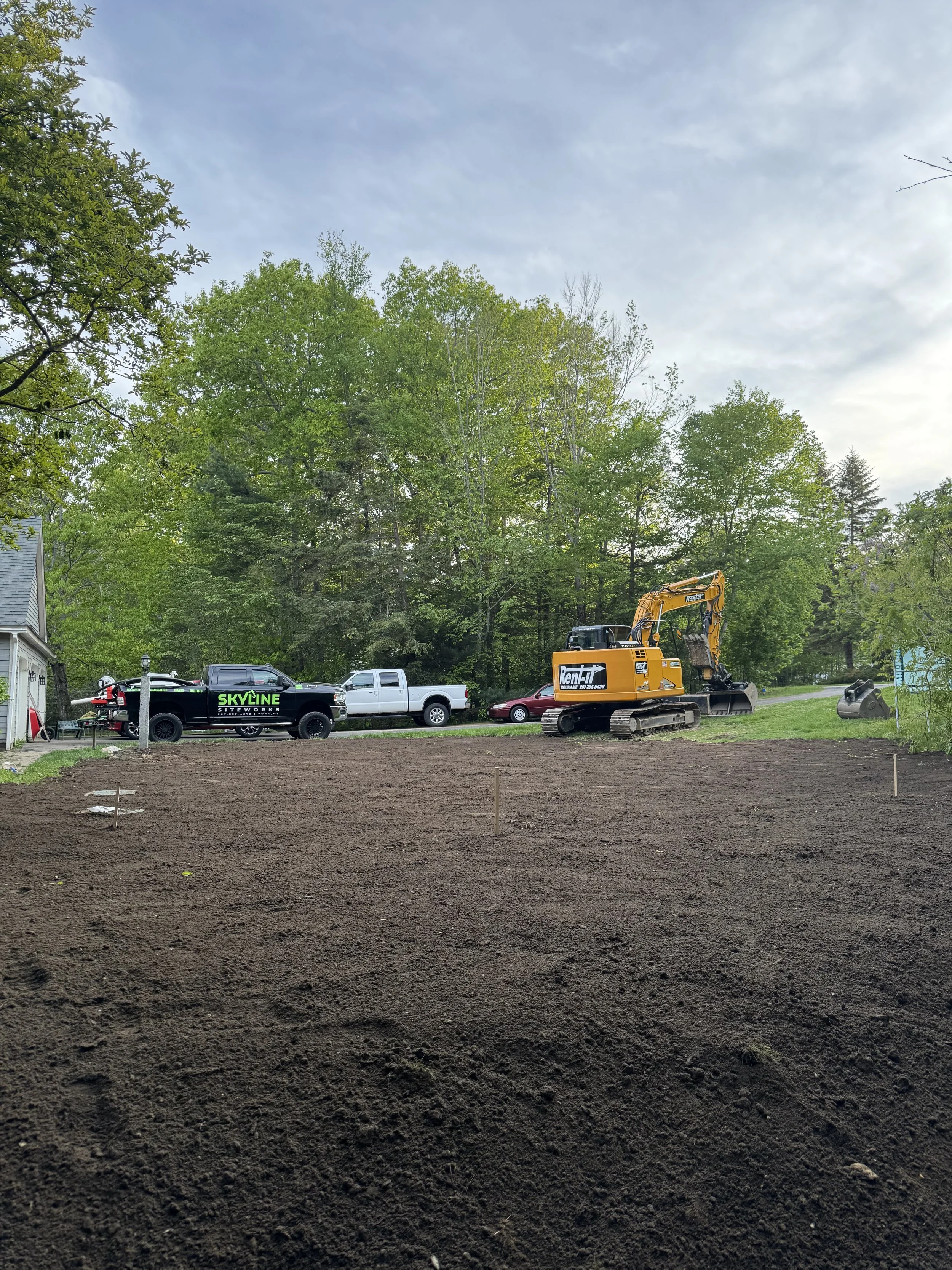 A construction site with excavator, trucks, and equipment, a freshly tilled soil area in the foreground, green trees in the background, and a partly cloudy sky.