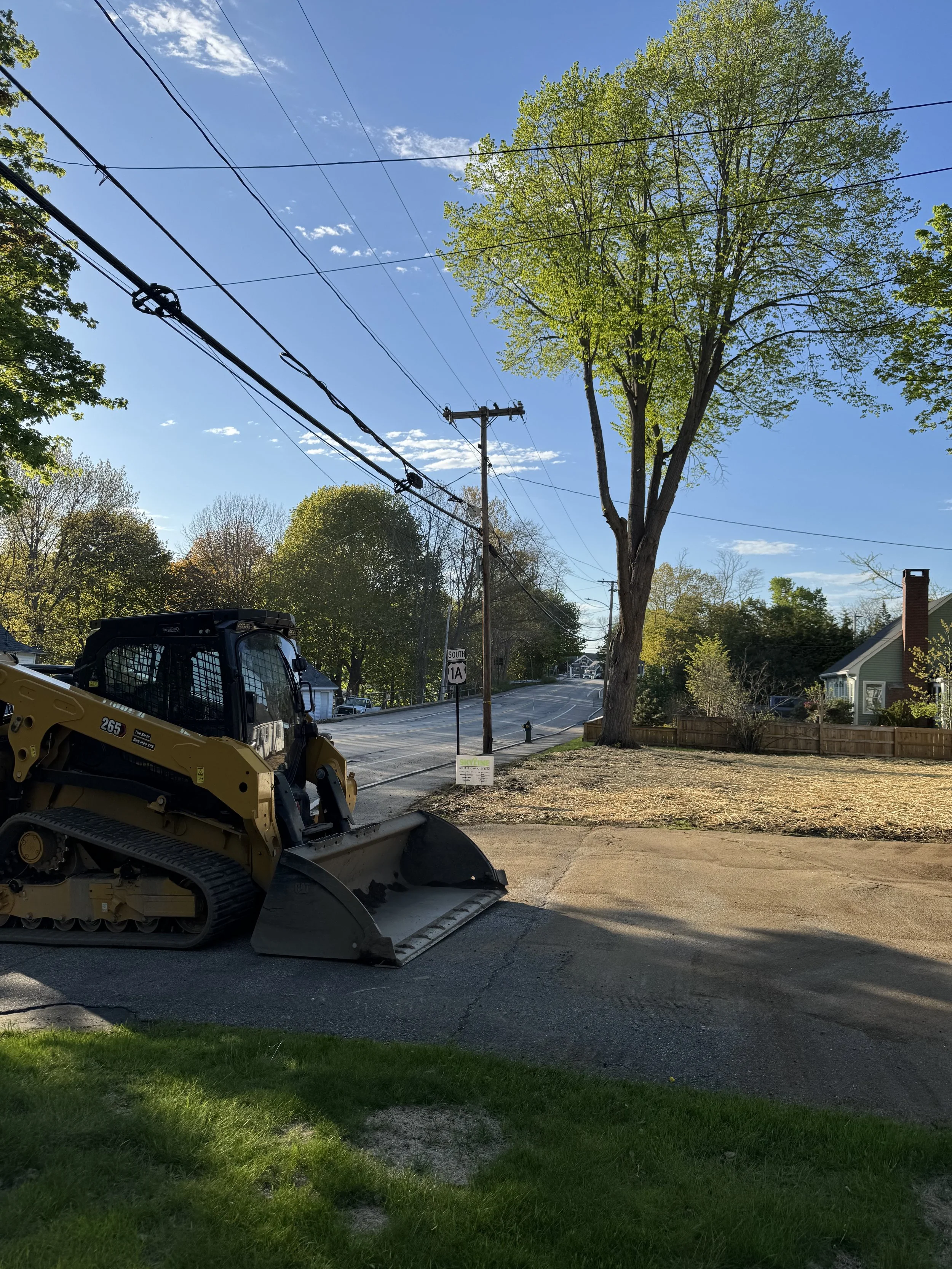 A construction vehicle parked on a residential street with trees, power lines, and houses, under a clear blue sky.