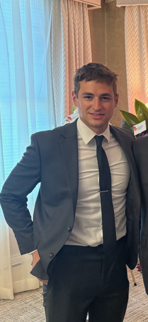 Young man in a navy suit and tie standing indoors near a window with curtains.