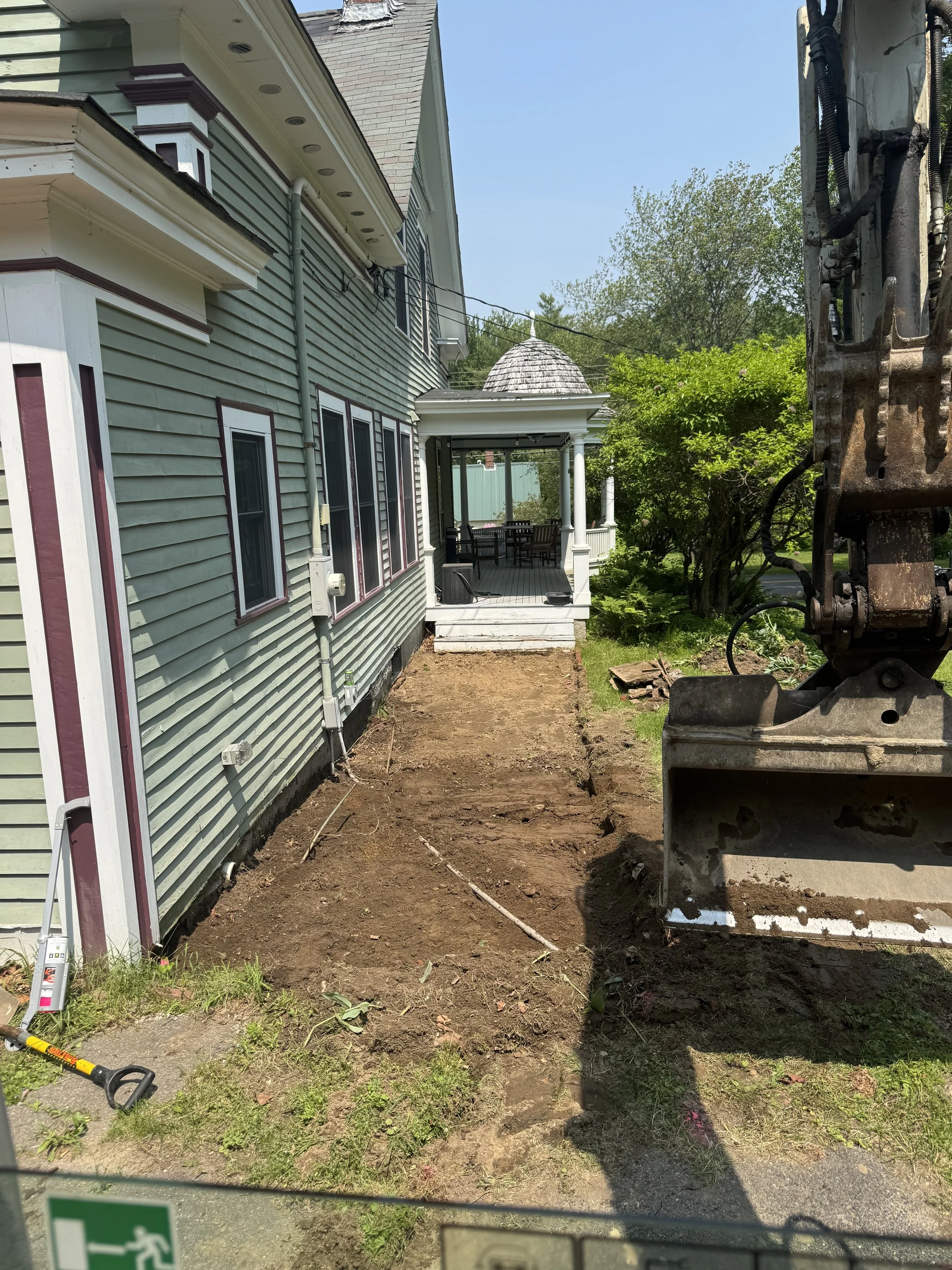 Side yard with dirt path next to a house under construction, with an excavator on the right and a porch with outdoor furniture in the background.