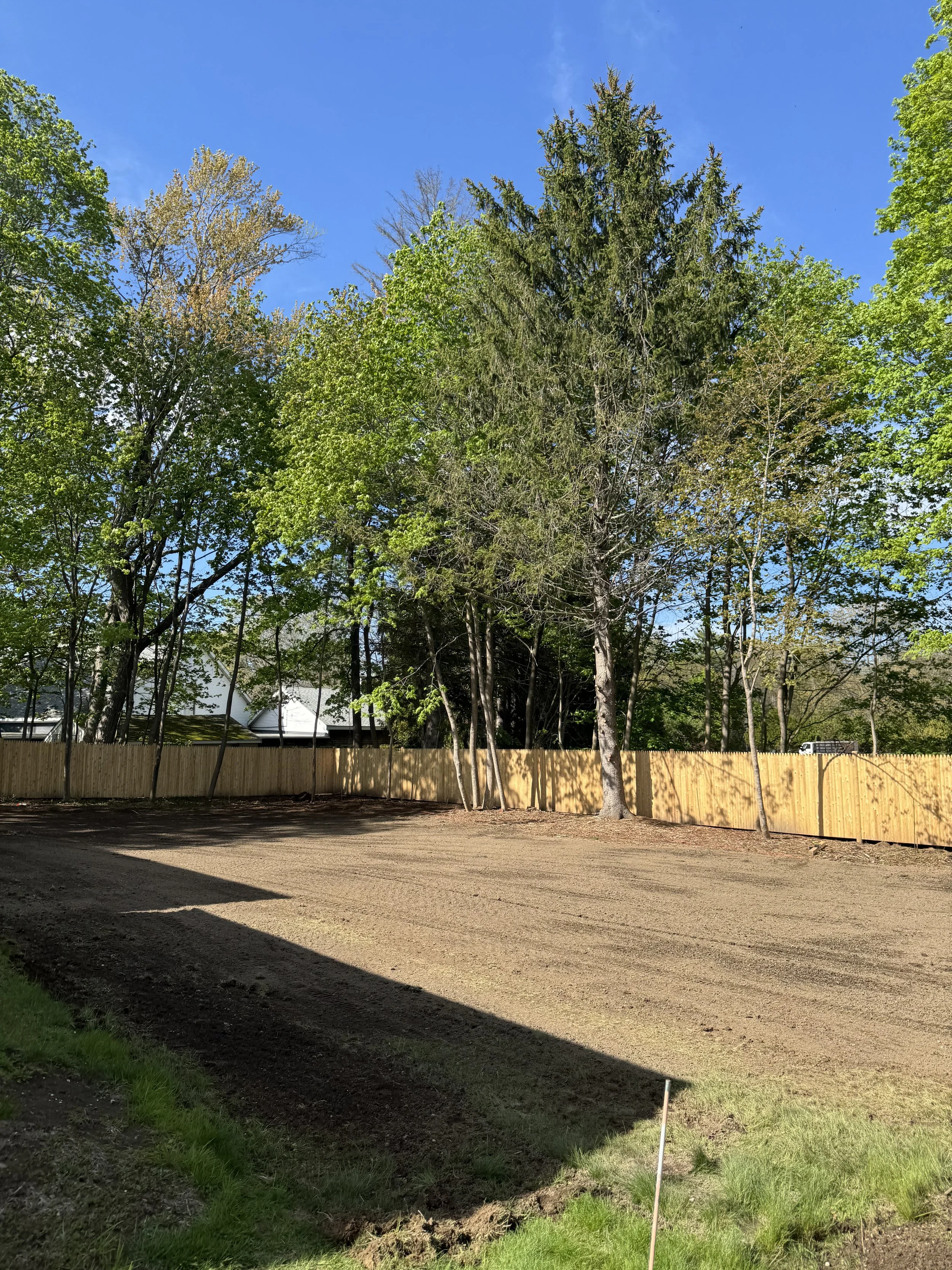 A cleared plot of land with a dirt surface, bordered by a yellow wooden fence. Behind the fence, there are several tall, leafy green trees and a house with a white roof, along with a wheelbarrow or similar equipment visible on the right side. The sky is clear and blue, indicating a sunny day.