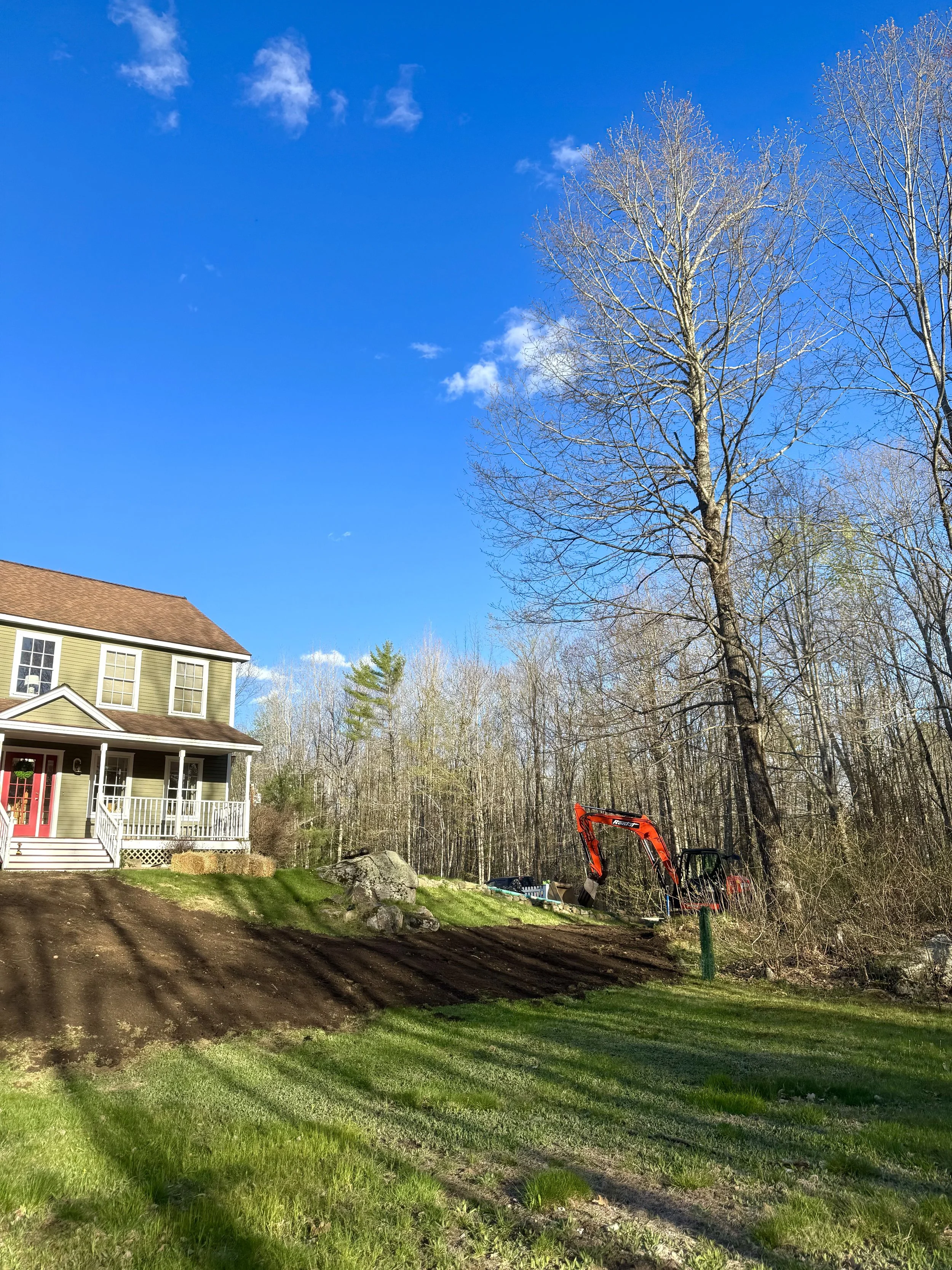 A house with a porch and windows on the left side, surrounded by a grassy yard and a dirt path. In the background, a forest with leafless trees and a clear blue sky with a few clouds. There is construction equipment, including a small orange excavator, on the right side of the image.