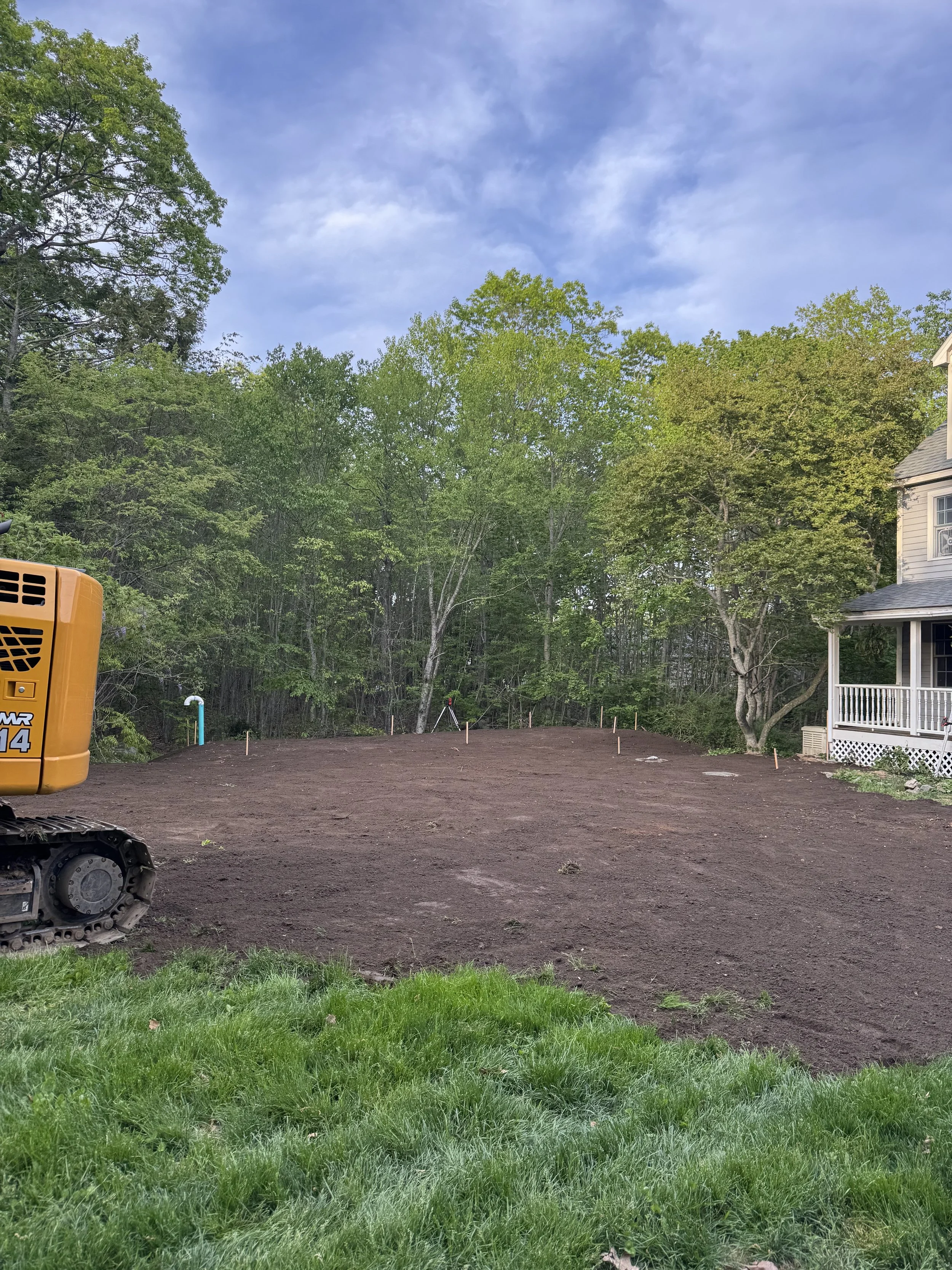 A yard with a patch of green grass in the foreground, a large area of freshly turned dirt, some small stakes, and a wooded area with trees in the background. Part of an orange construction vehicle is visible on the left side. A house with a porch is on the right side.