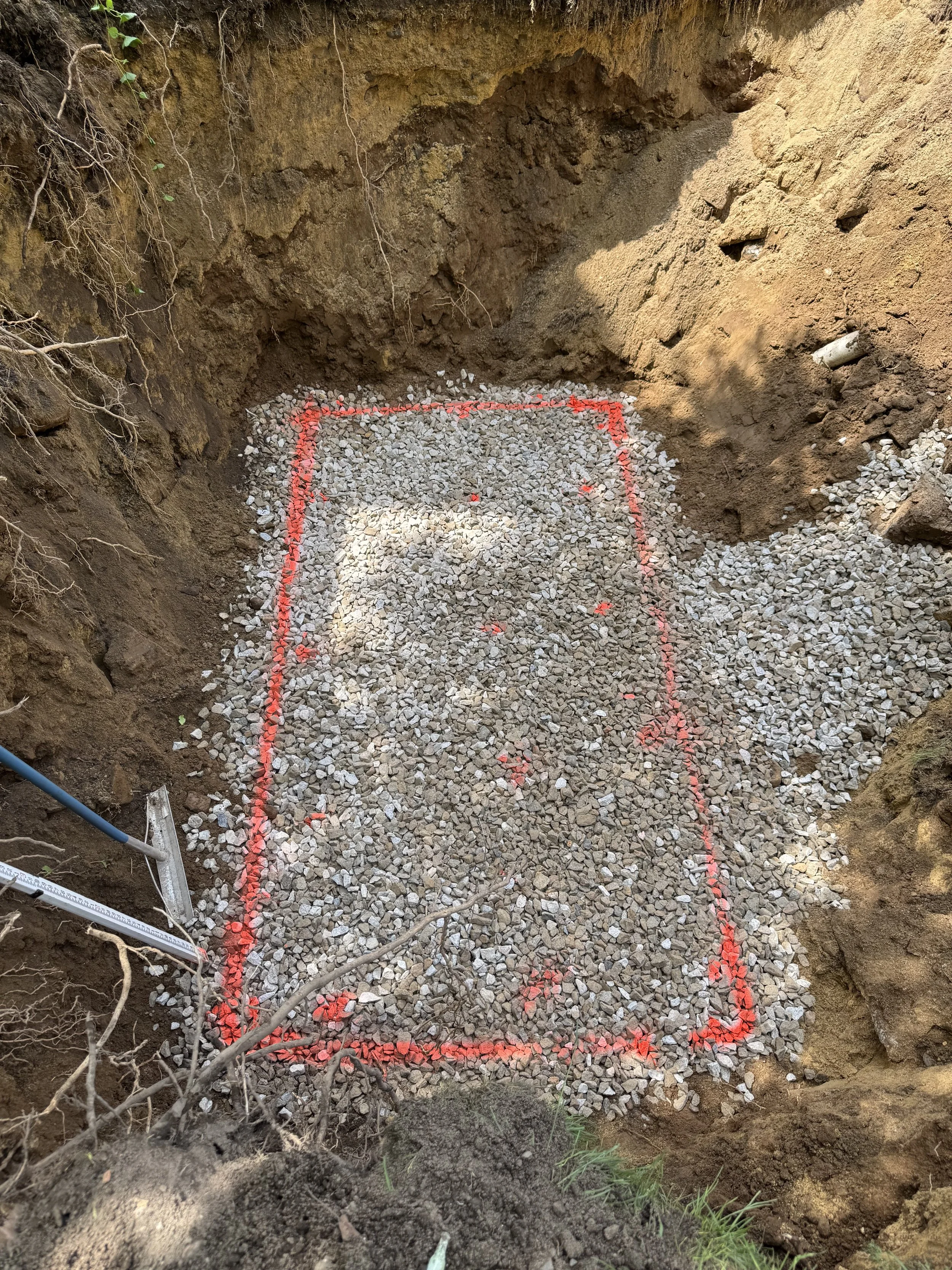 A rectangular area outlined with red markings in an excavation site filled with small gravel stones. The surrounding soil is excavated, revealing tree roots and soil layers.