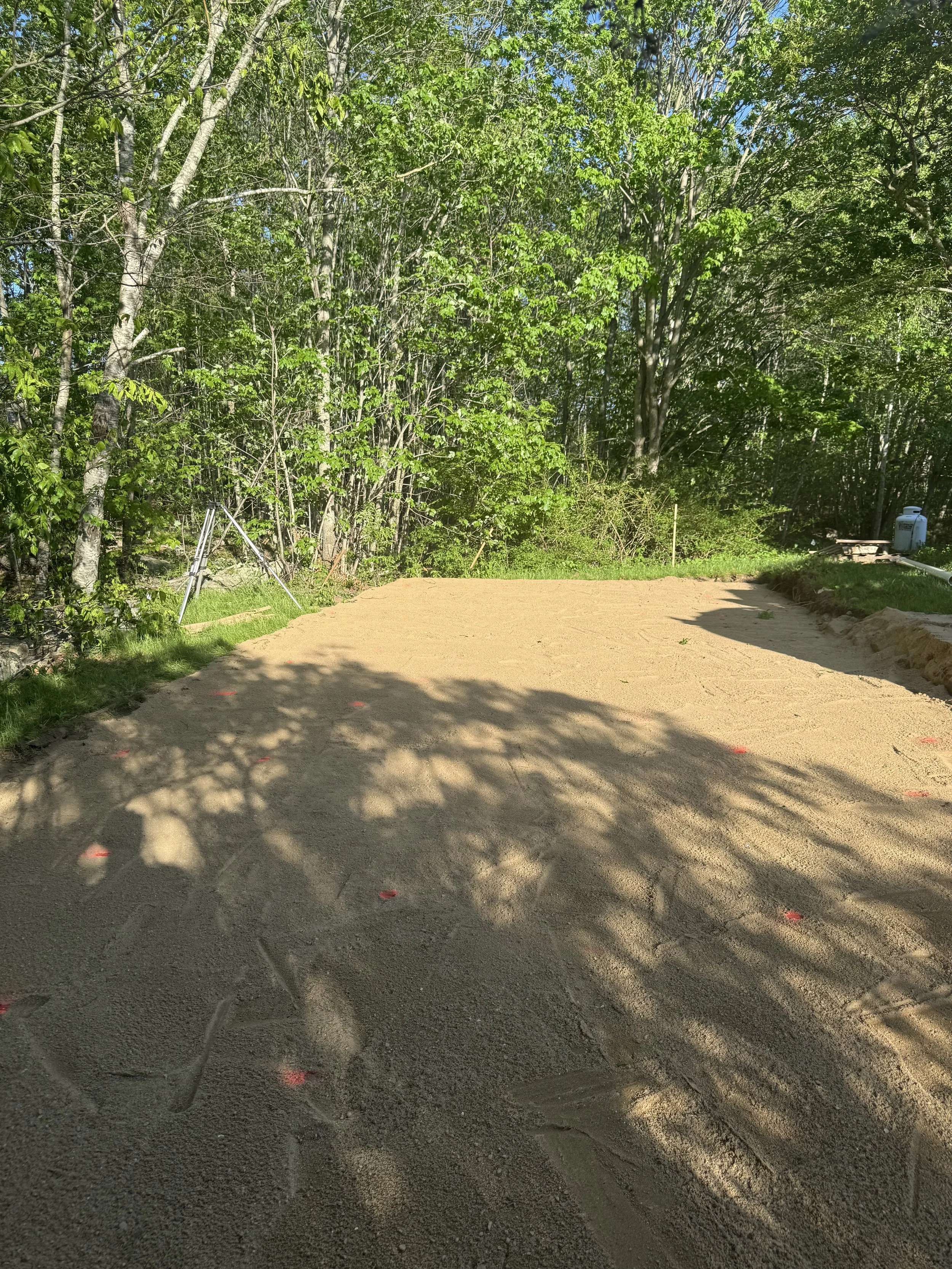 A construction site with a partially leveled dirt surface, surrounded by green trees and bushes, with some construction equipment and materials visible in the background.
