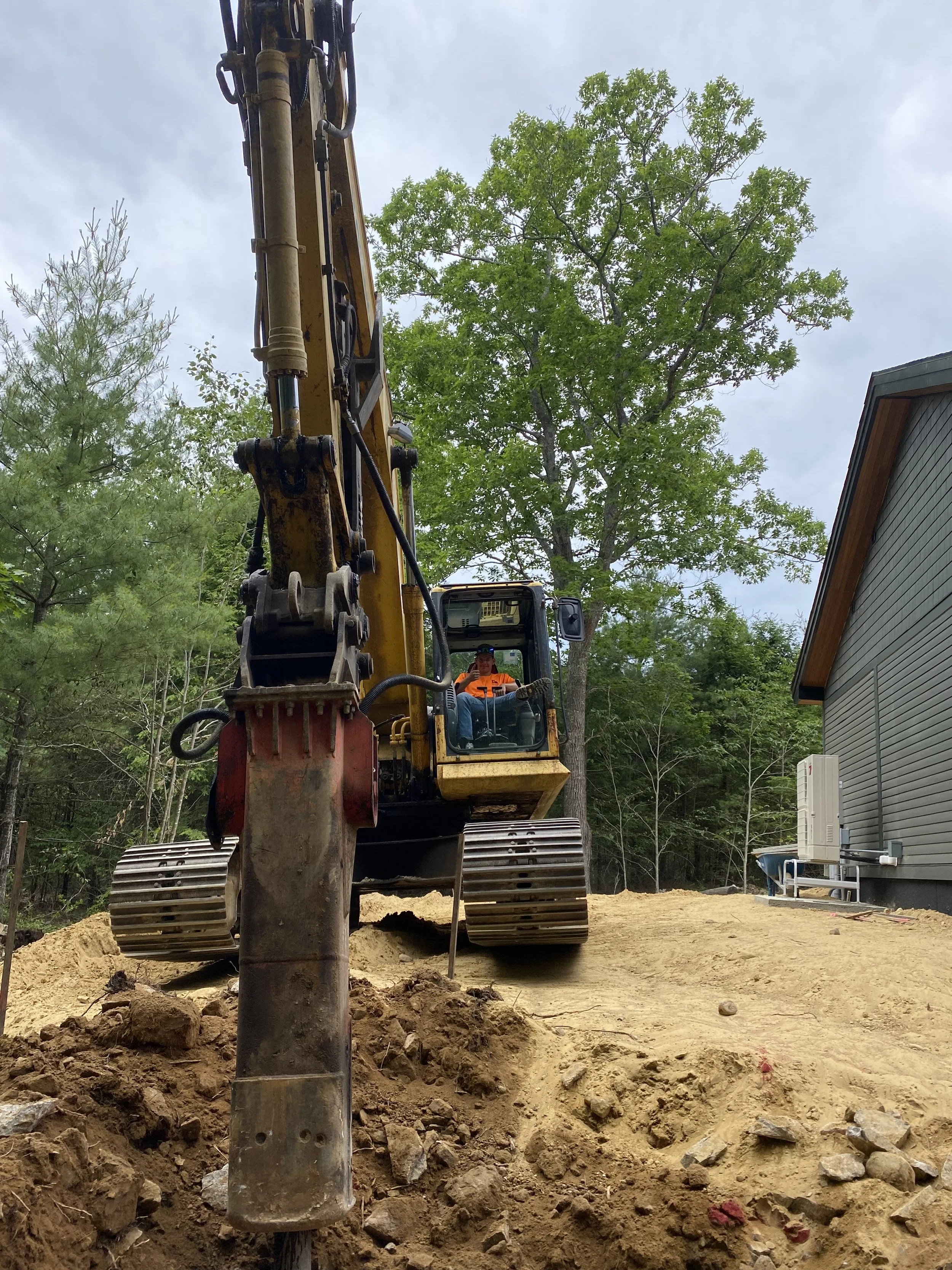 A construction worker operating a yellow excavator on a dirt construction site next to a gray building with trees in the background.