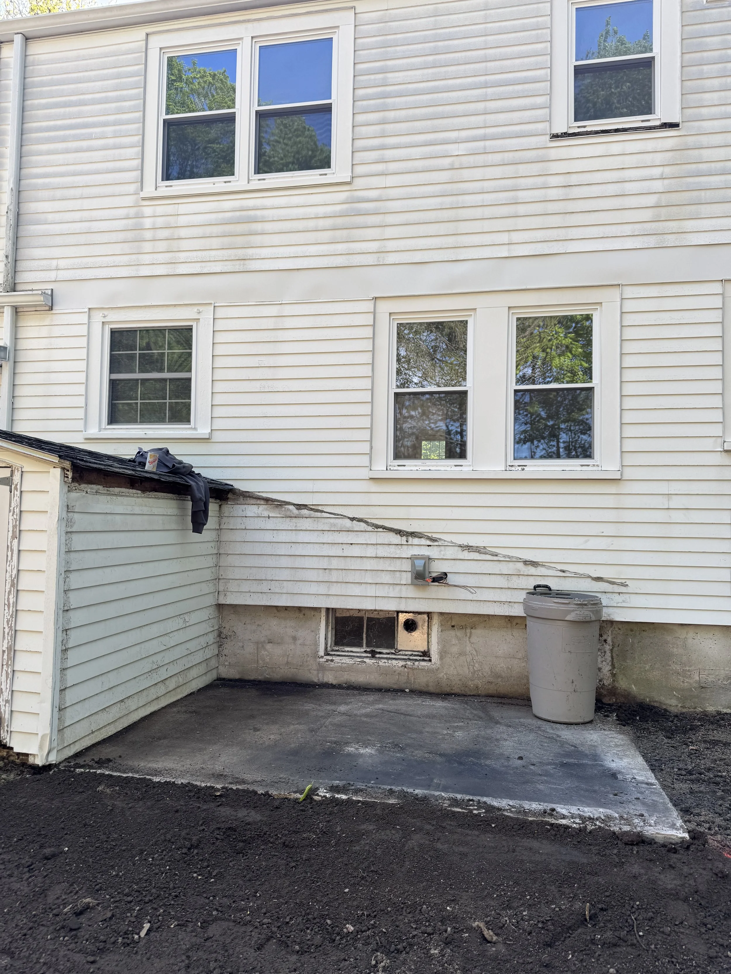 The back of a house with three white-framed windows, some outdoor hardware, and a small section of dark asphalt or concrete ground with a large gray trash can.