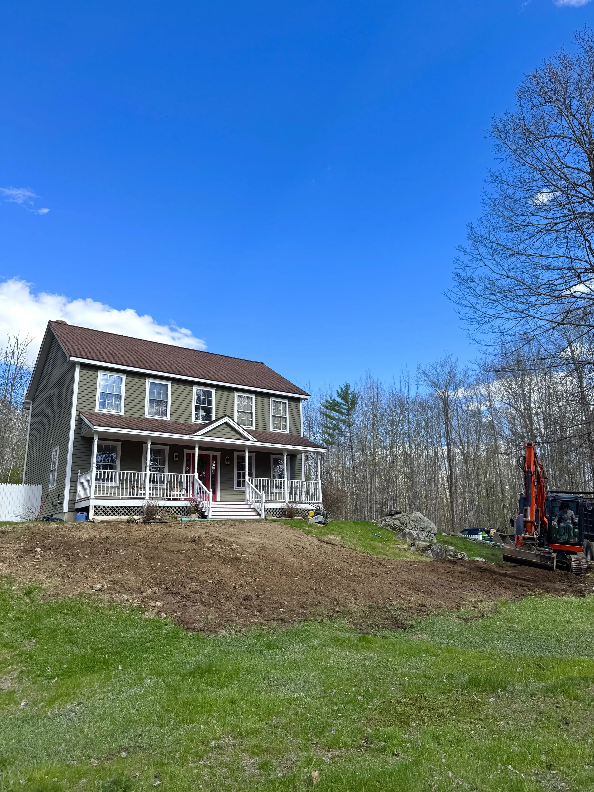 A house with beige siding, white trim, and a dark red roof is under construction or renovation on a grassy lot with patches of dirt. There is a small porch with white railings. A backhoe is on the right side of the image, and there are leafless trees and a bright blue sky in the background.