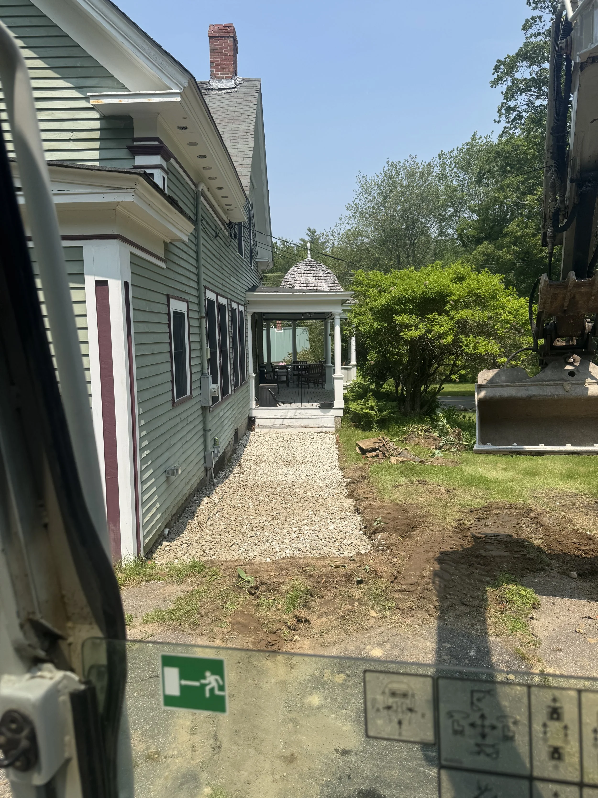 A green house with a porch and outdoor dining area, being landscaped with a gravel path and some soil dug up, visible from inside a construction vehicle.