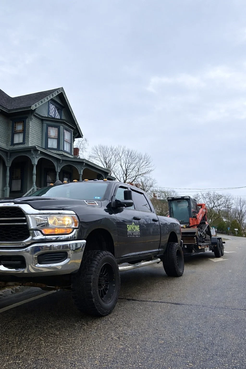 A black pickup truck with a logo and phone number on the door, towing construction equipment on a flatbed trailer, parked on a street in front of a Victorian-style house with blue-green paint and intricate woodwork