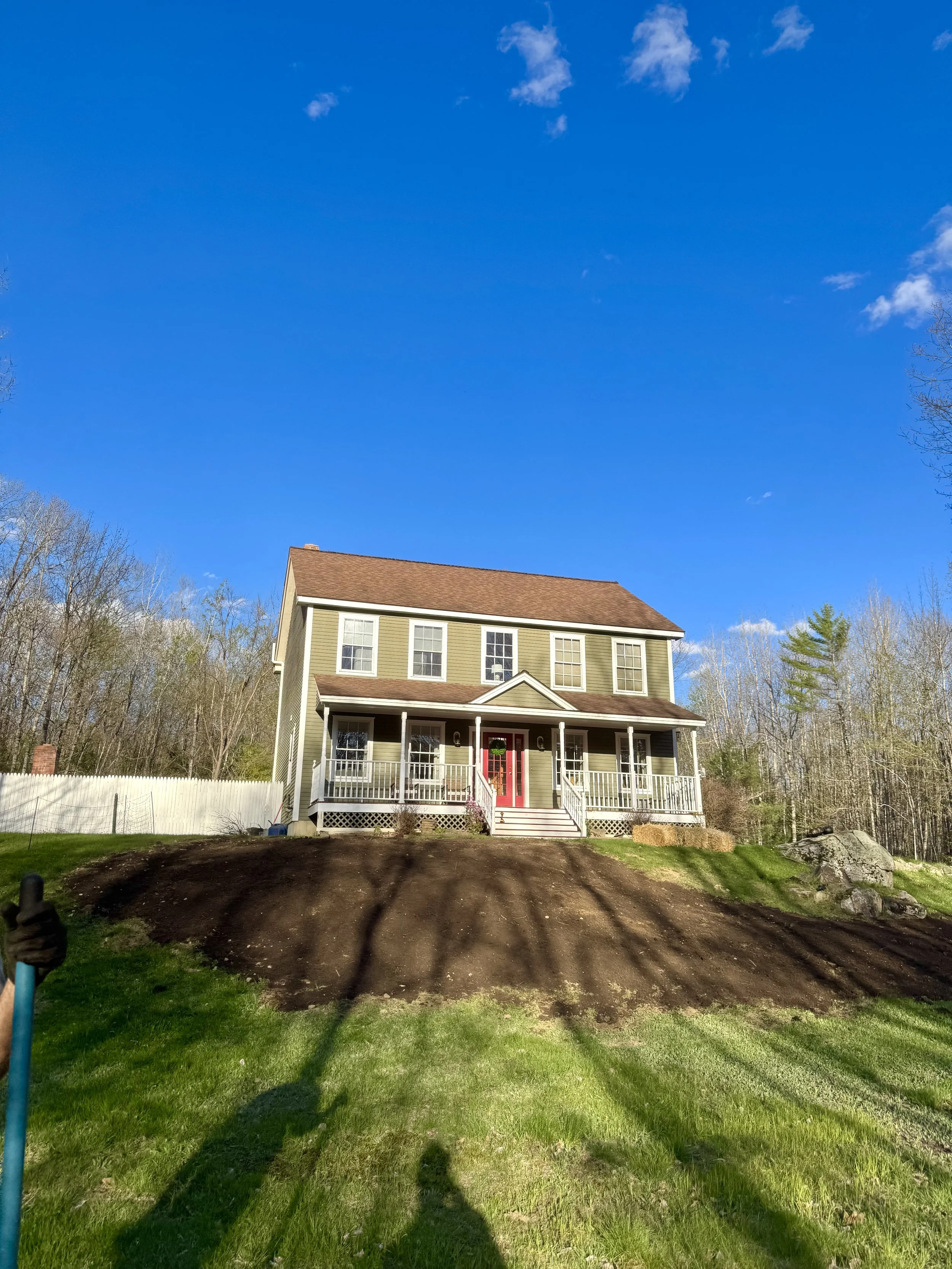 A two-story house with a beige exterior, brown roof, white trim, and a red front door, surrounded by a grassy yard and trees under a blue sky with some clouds.