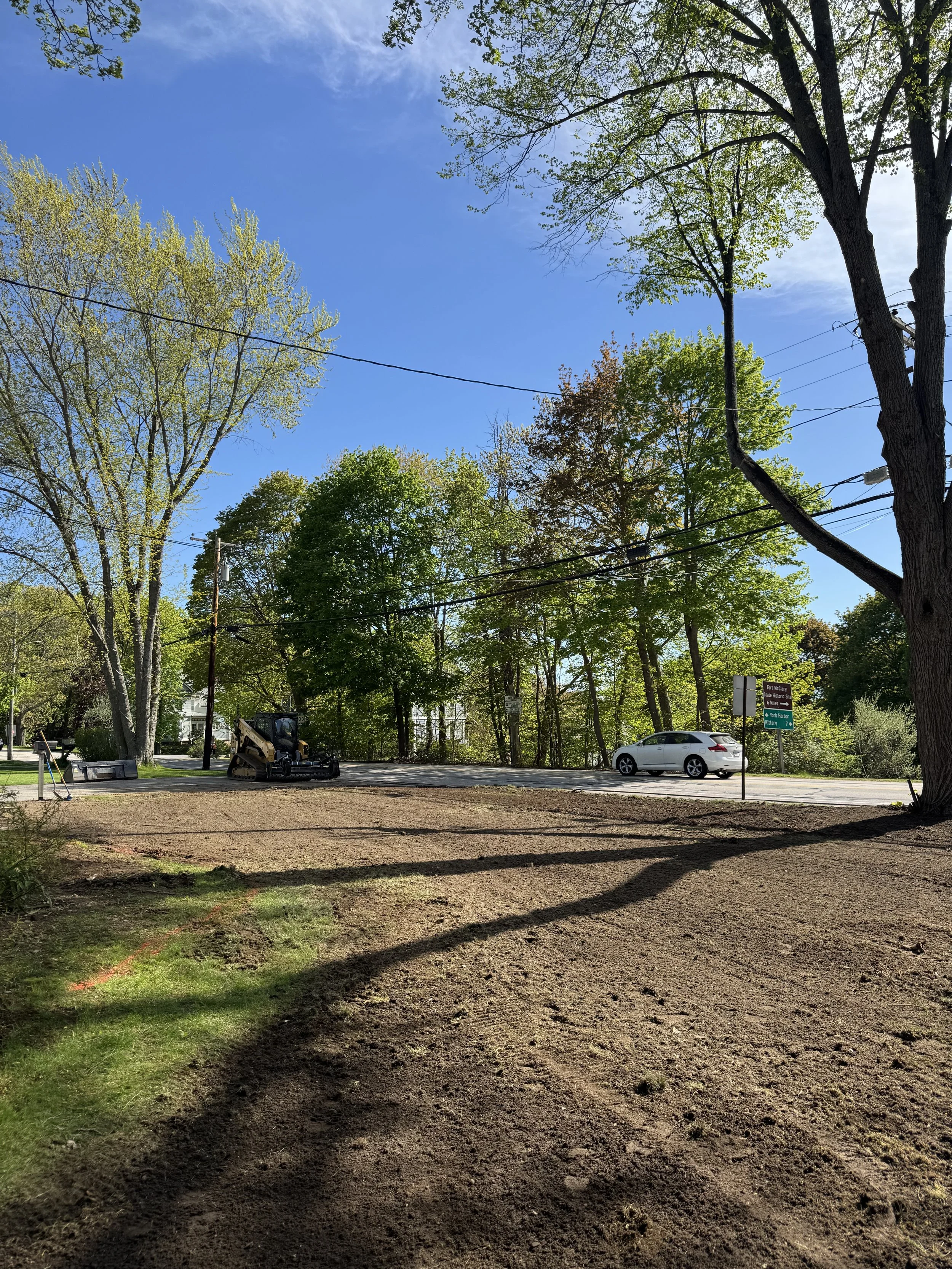 A backyard scene with trees, a patch of dirt, and a parked white car near a road. There is a construction machine and signs on the street, with power lines overhead.