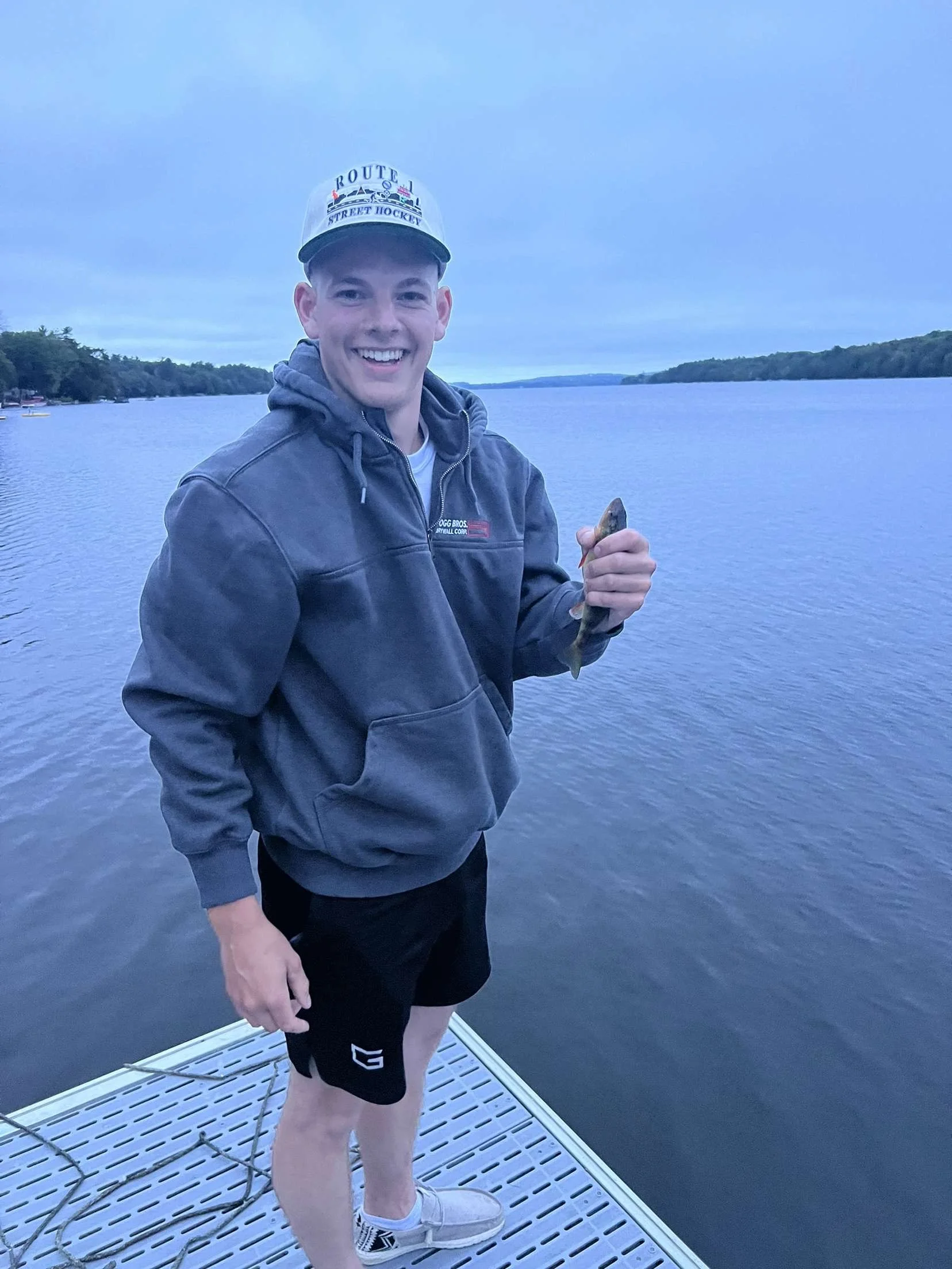 A young man standing on a dock, holding a small fish, smiling, with a lake and trees in the background, wearing a hoodie, shorts, and a baseball cap.