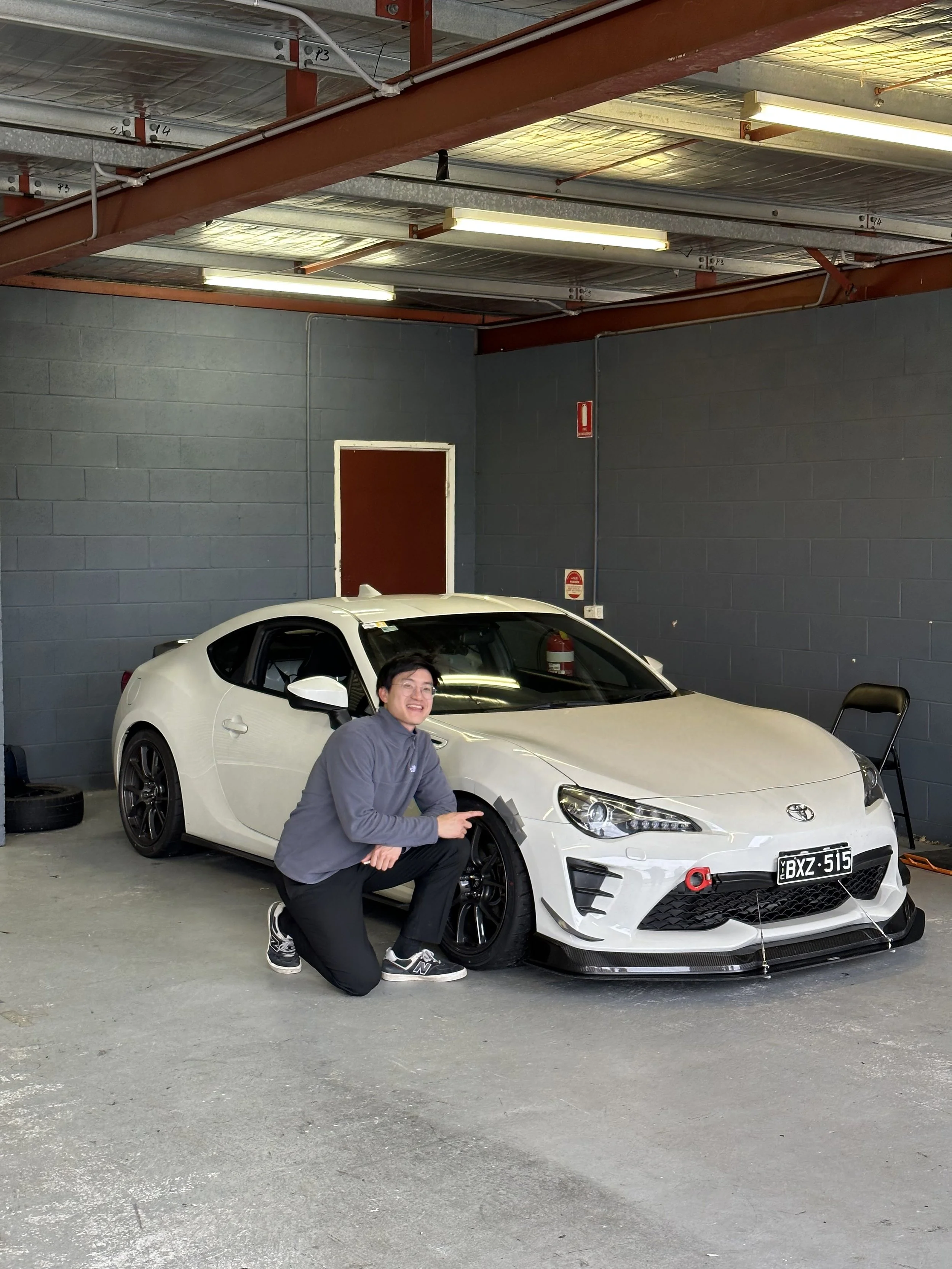 A man kneeling next to a white sports car in a garage, pointing at the front wheel, smiling. The garage has a gray brick wall, fluorescent lights, a backpack, a chair, and a fire extinguisher.