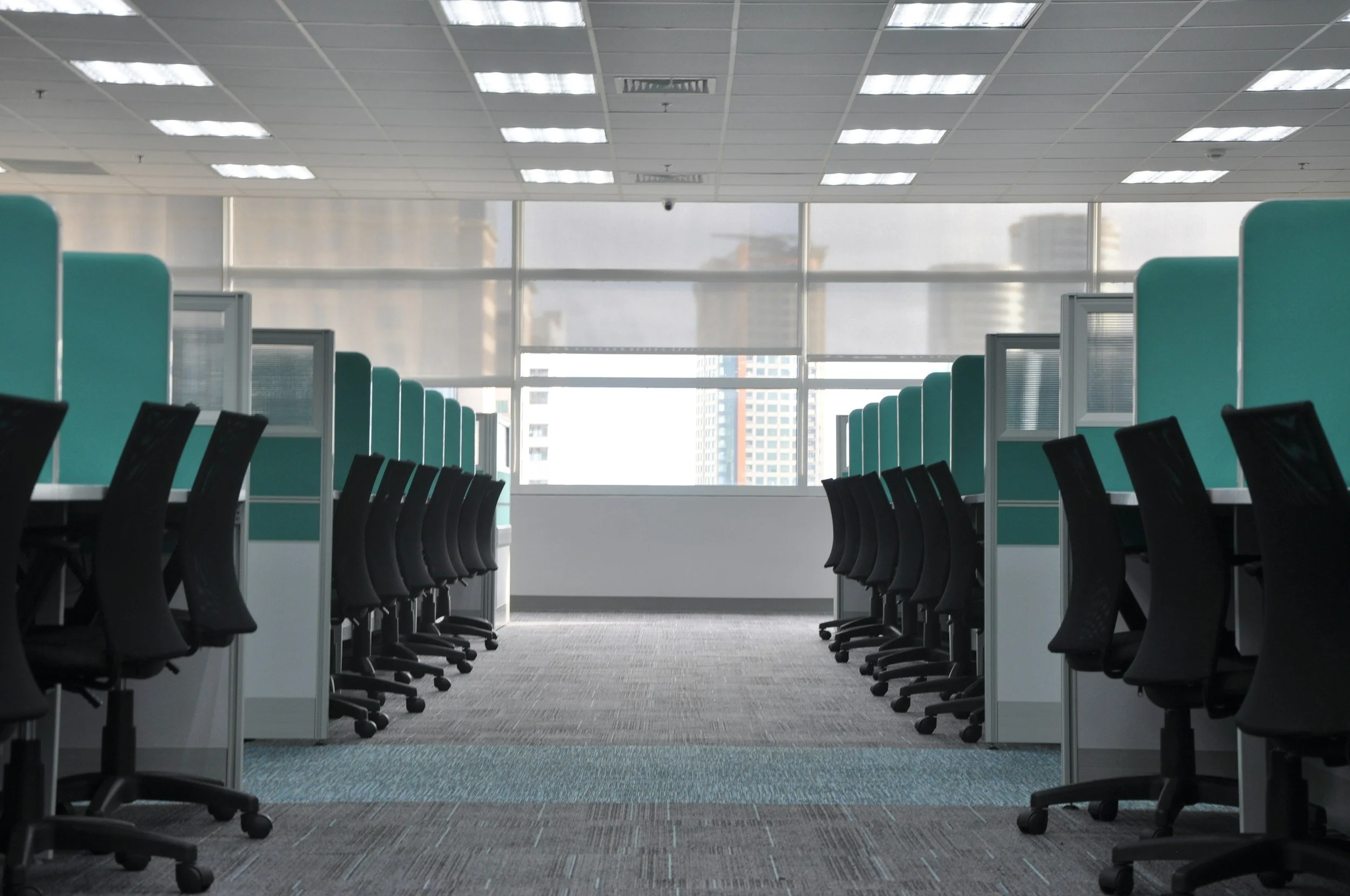 Empty office space with rows of black chairs, cubicles with teal dividers, large windows, and cityscape view, illuminated by ceiling lights.