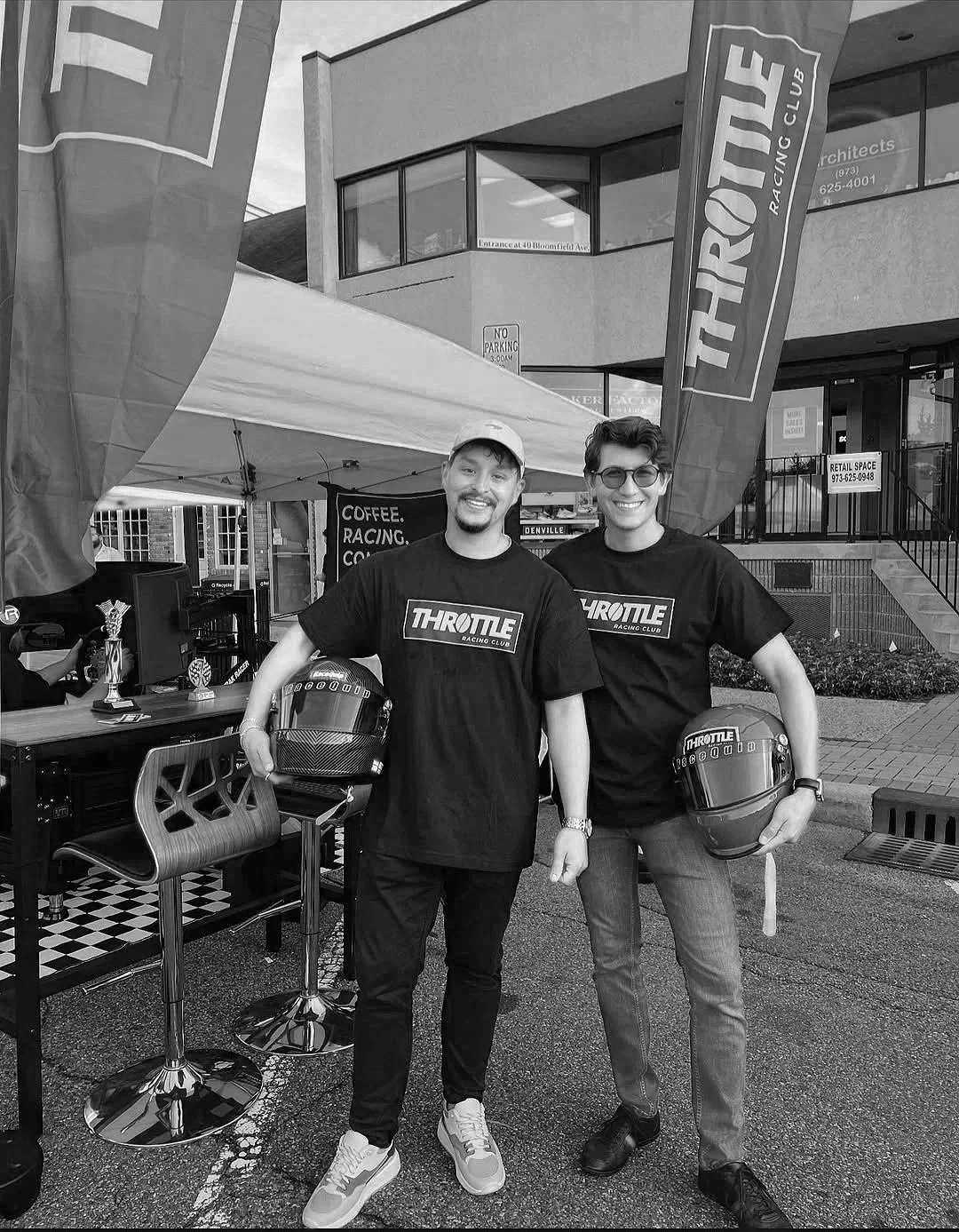 Two young men wearing black THROTTLE Racing Club t-shirts, standing outdoors with one holding a motorcycle helmet in each hand, smiling and posing for the photo. There are flags and a tent behind them, with a building in the background.