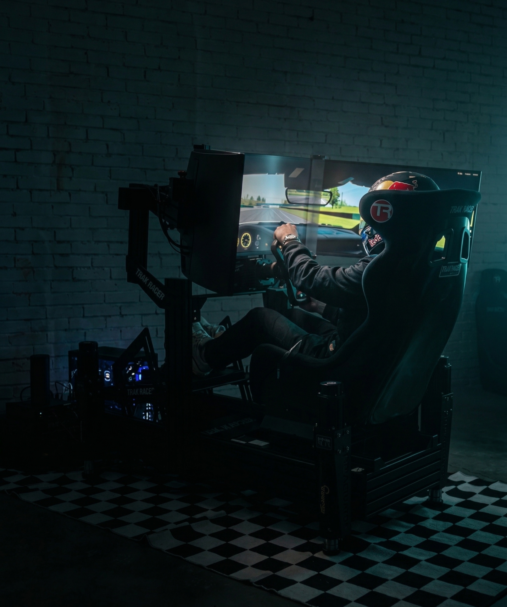 Person in a racing simulator seat wearing a helmet, operating a high-tech driving system with multiple screens, set against a dark room with a brick wall.