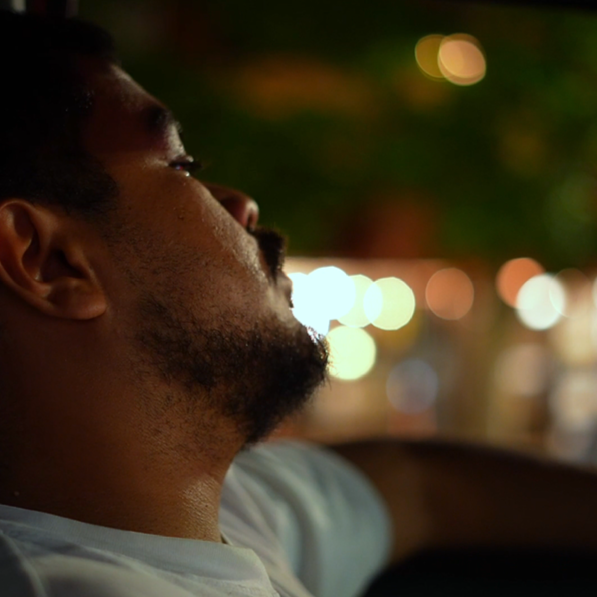 A man with short hair and a beard is sitting in a vehicle at night, looking out with city lights blurred in the background.