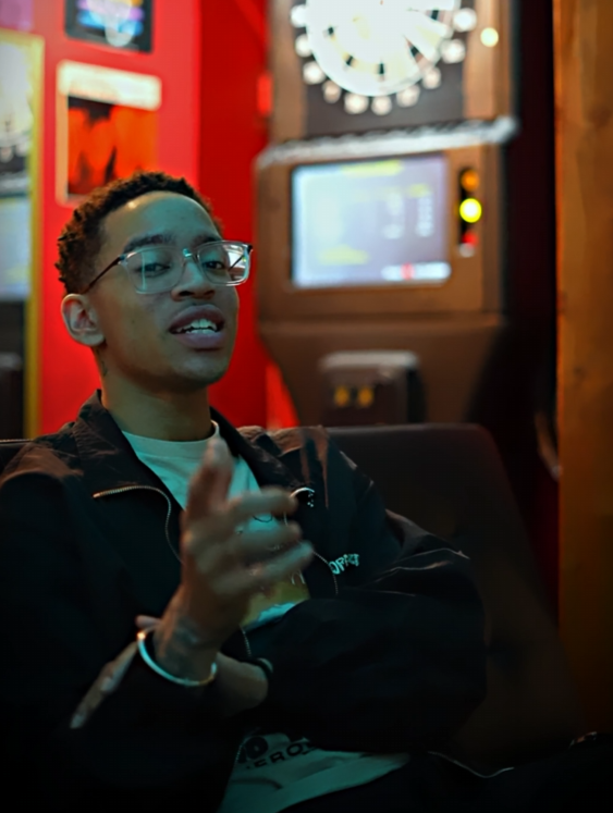 Young man wearing glasses, sitting in a bar or arcade, pointing at the camera with a dartboard and gaming machine in the background.