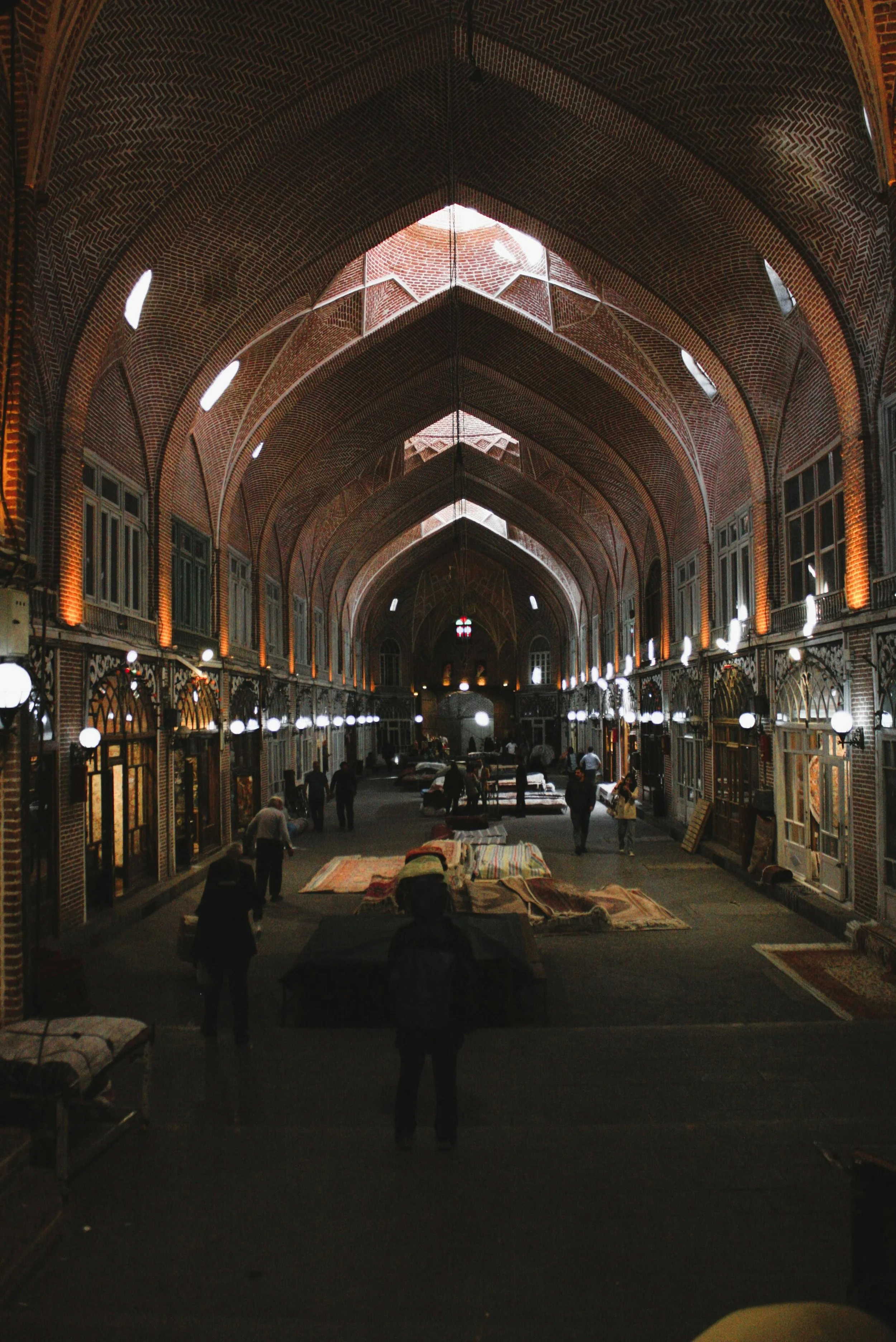 Interior view of a spacious, arched market with brick walls and ceiling, illuminated by hanging lights, with vendors displaying rugs and textiles.