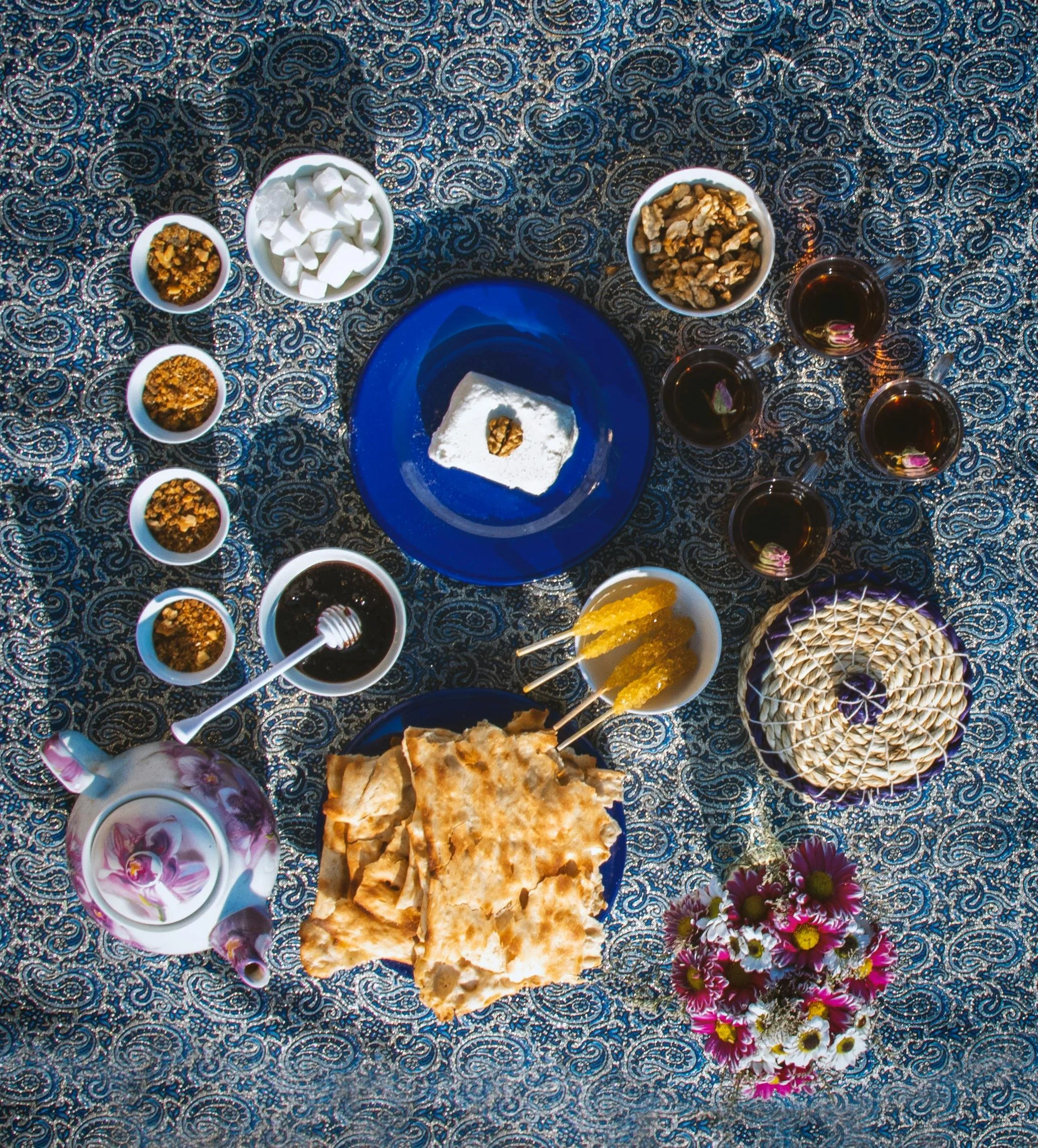 A traditional Middle Eastern meal spread on a patterned tablecloth. The spread includes a piece of white cheese with a walnut on top, various bowls of honey, nuts, and spices, a teapot, glasses of tea with flowers, a plate of flatbread, a basket of a decorative bread, and a small bouquet of pink and white flowers.