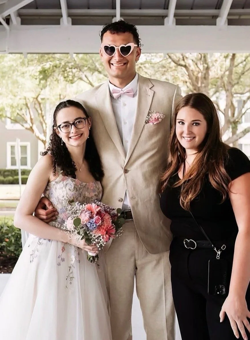 A bride and bridesmaids hold large bouquets of flowers over their faces during an outdoor wedding, with trees and pink blossoms in the background.