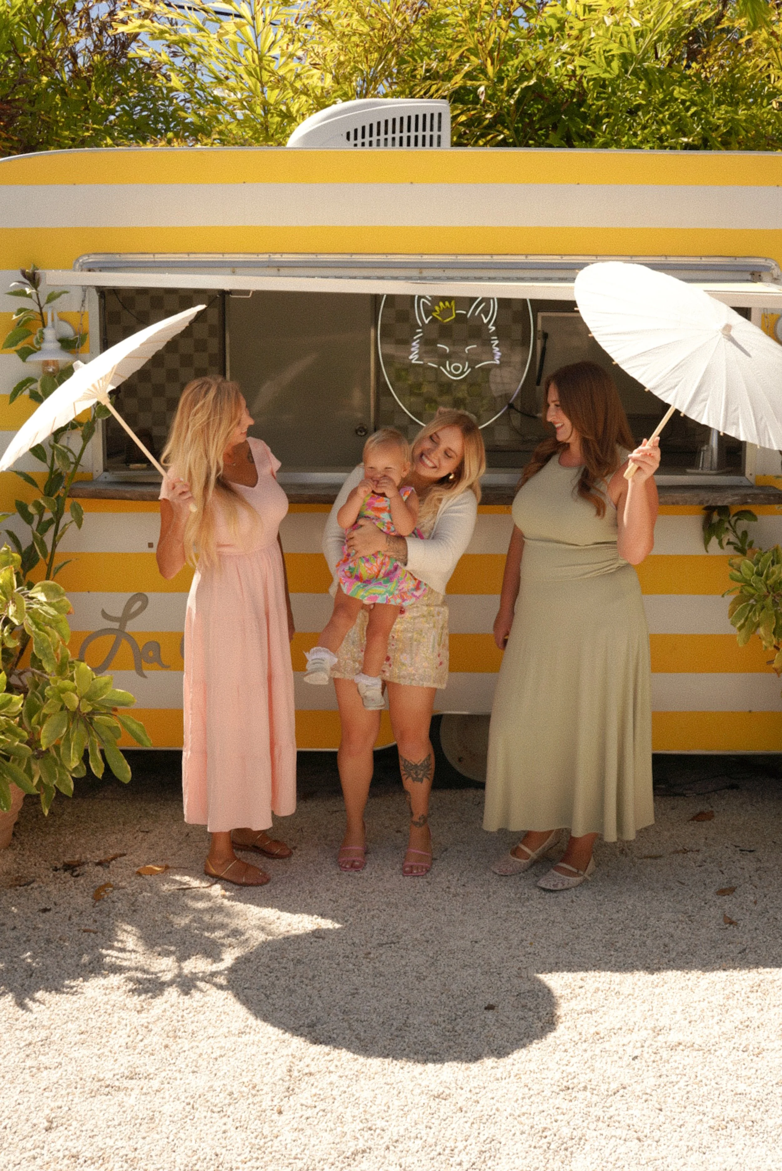 Four women and a young girl standing in front of a yellow and white striped food truck, holding white umbrellas, smiling and enjoying the sunny day.
