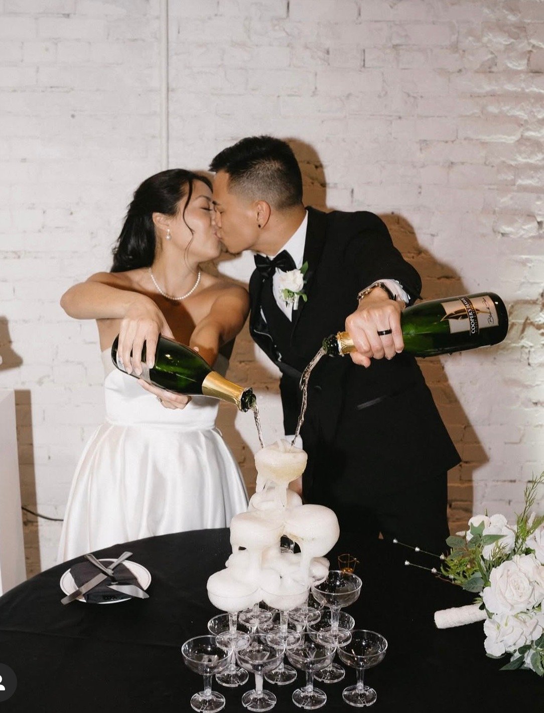 A bride and groom in wedding attire pour champagne into a champagne tower at their wedding reception.