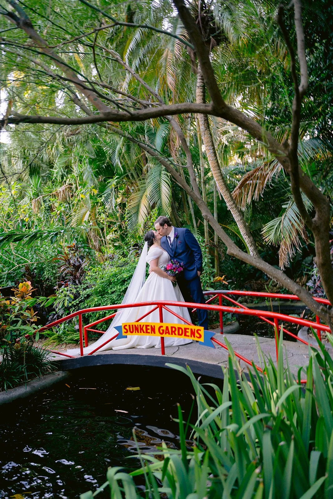 A couple sharing a kiss at their outdoor wedding ceremony, surrounded by bridesmaids in teal dresses holding bouquets, with trees and sunlight in the background.