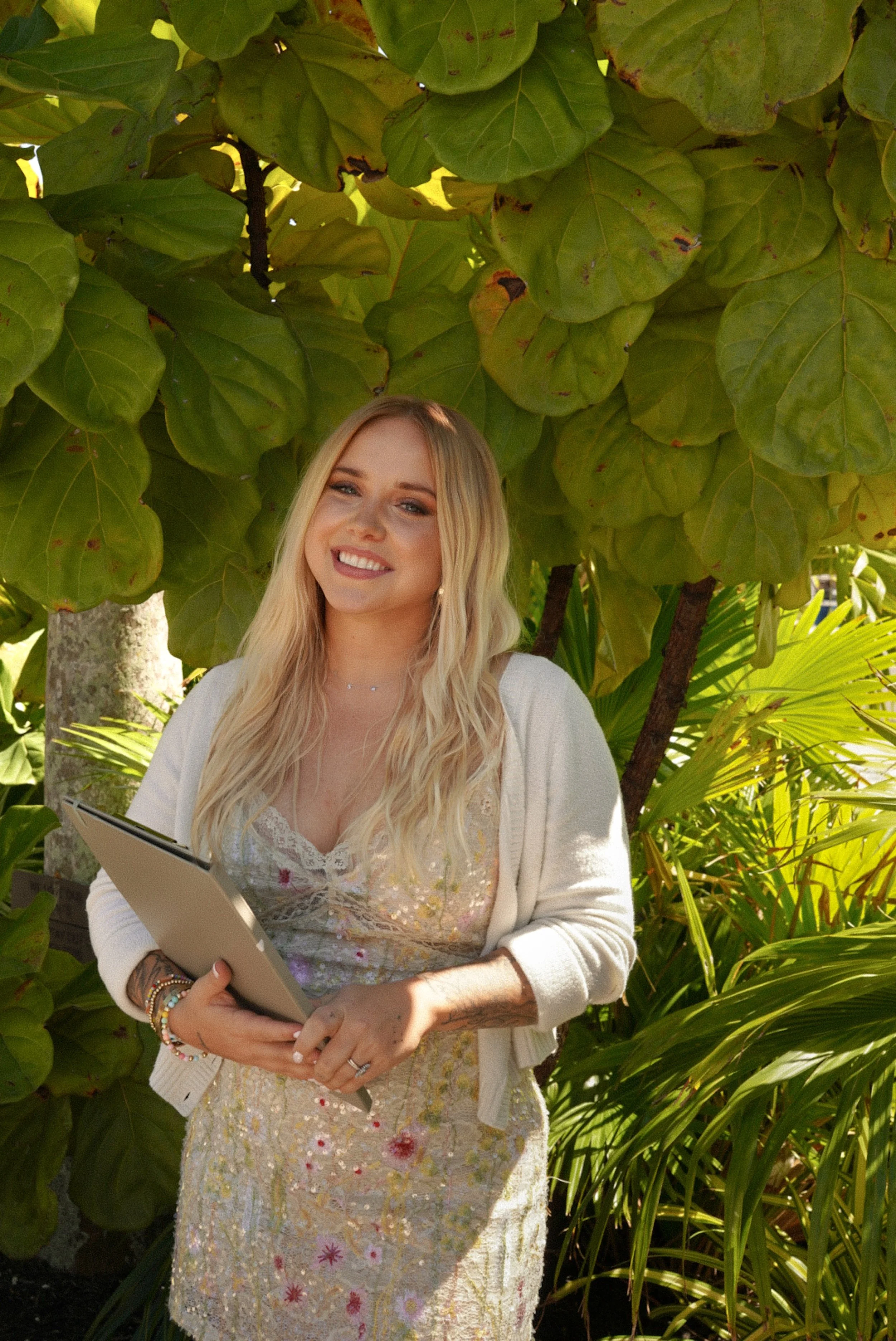 A young woman with long blonde hair smiling and holding a beige notebook outdoors amidst green foliage and trees.