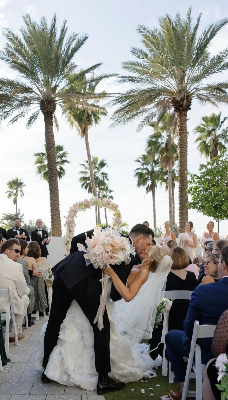 A bride and bridesmaids hold large bouquets of flowers over their faces during an outdoor wedding, with trees and pink blossoms in the background.
