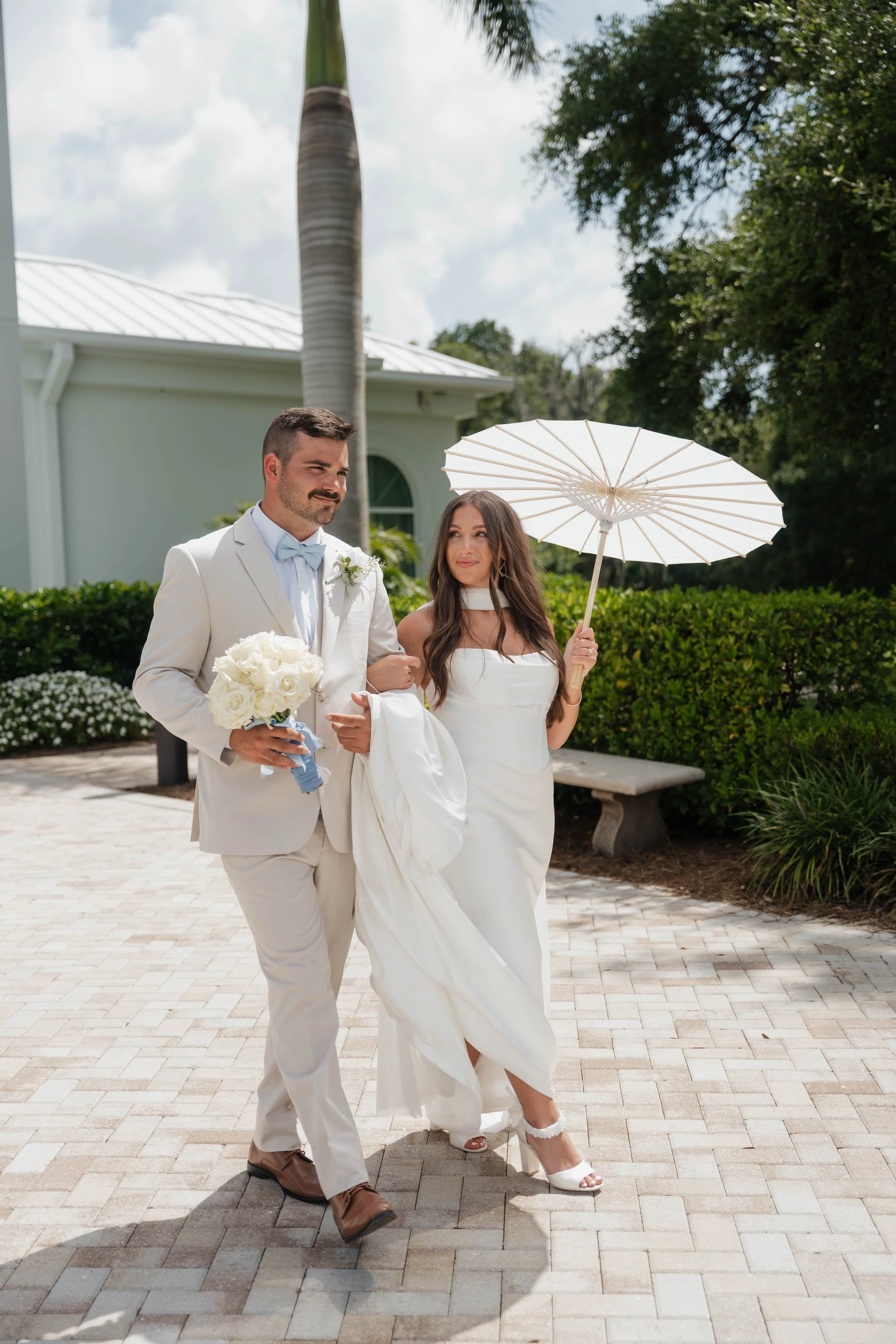 A bride and groom walking outdoors on a sunny day, with the bride holding a white parasol. The groom is dressed in a light-colored suit and holding a bouquet of white flowers. The bride wears a white wedding dress and heel sandals, with long dark hair.