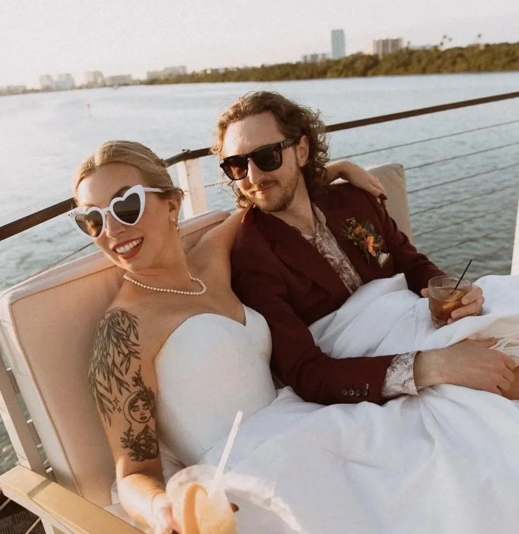 A woman and a man are sitting together on a boat, smiling, with water and a city skyline in the background. The woman is wearing heart-shaped sunglasses, and the man is wearing dark sunglasses. The woman is dressed in a white strapless dress, and the man is wearing a maroon blazer. They are enjoying drinks, and it is sunset.